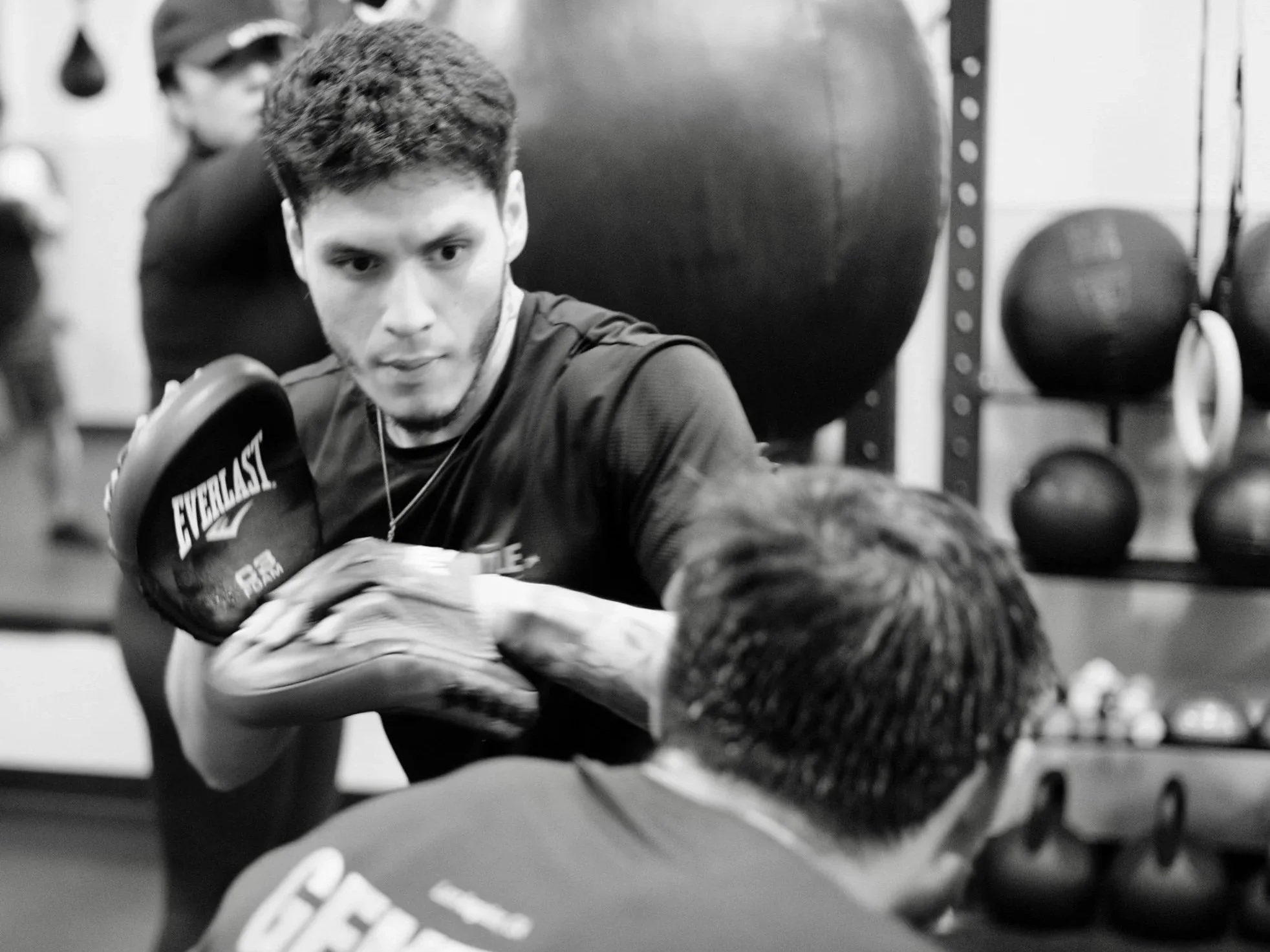 A young man with dark hair and a beard sparring in a boxing gym, wearing boxing gloves, with other gym equipment like medicine balls in the background.