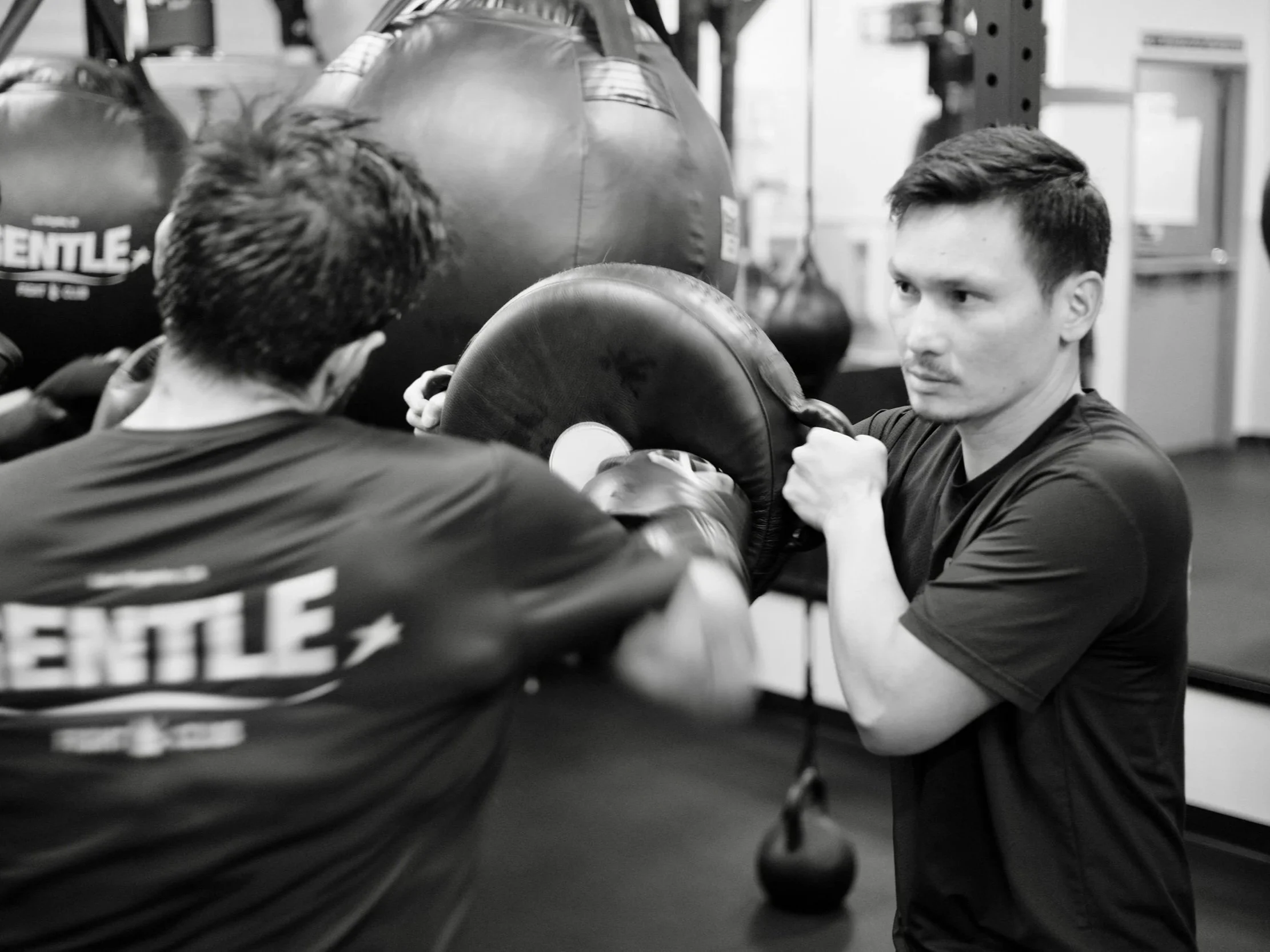 Two men with short dark hair and athletic build training with boxing pads in a gym. One man, with a serious expression, holds the pads while the other prepares to punch. Boxing gloves and equipment are visible, with a large black punching bag in the 