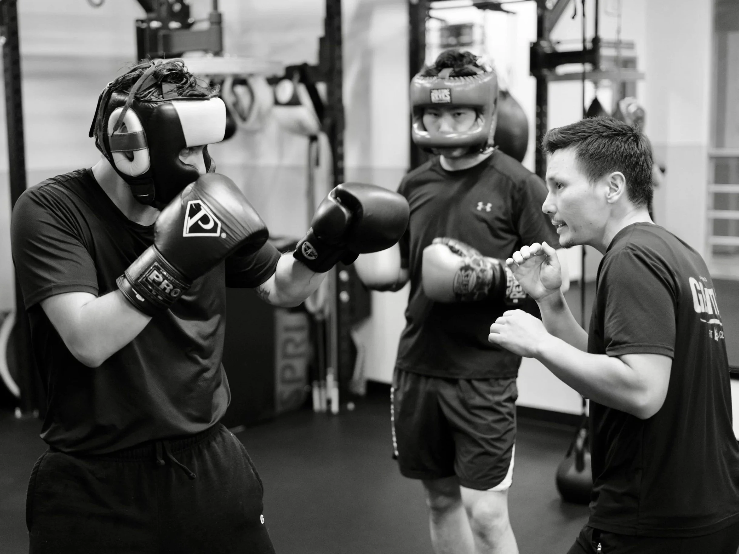 Two young men wearing boxing gloves and protective headgear sparring in a gym. A third young man with boxing gloves stands between them, possibly coaching or instructing. The scene is in a martial arts or boxing training facility.