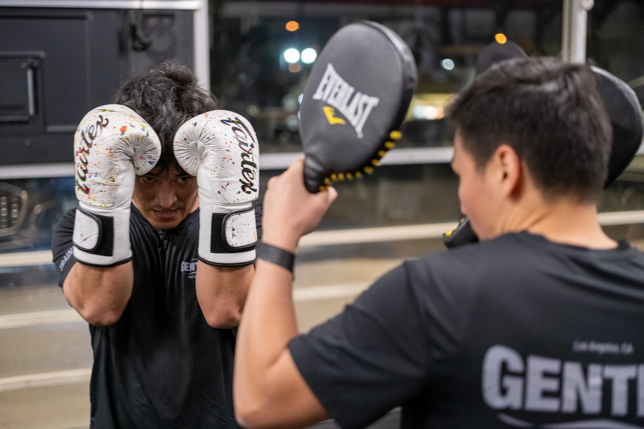 Two men in boxing gloves and athletic clothing practicing boxing in a gym at night, with the man on the left holding his gloves up in a defensive stance and the man on the right holding focus mitts for training.