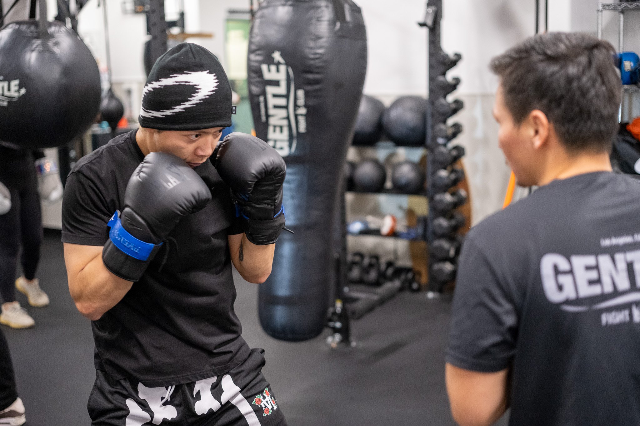 Two men are in a boxing gym, practicing boxing. One man is wearing a black beanie and black boxing gloves, facing the other man who is wearing a black shirt with text on the back. The gym has boxing equipment including hanging black punching bags, shelves with black medicine balls, and other training gear.