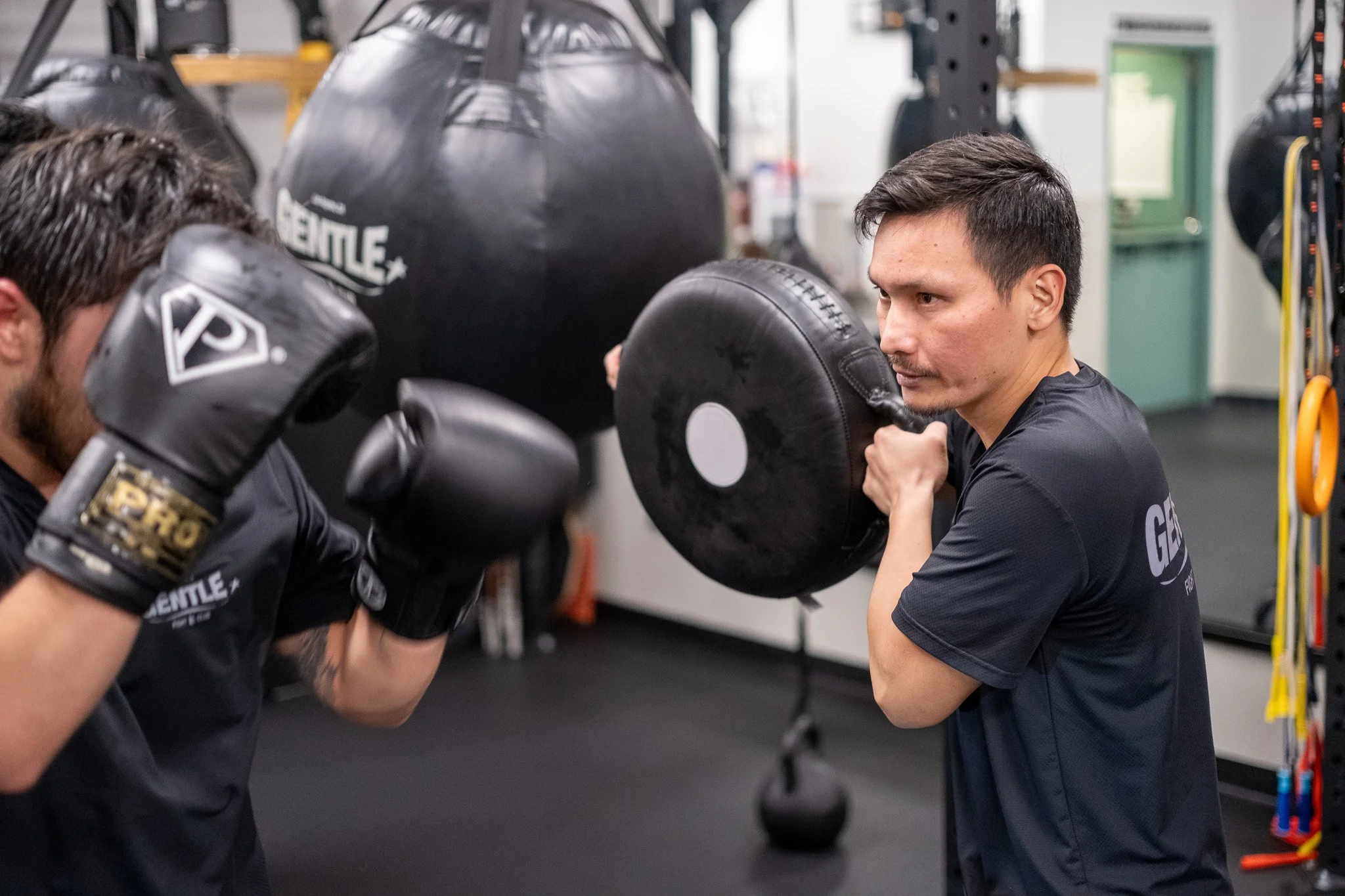 A man is coaching a woman in boxing, with both standing in front of a speed bag in a gym. The woman is wearing boxing gloves and headgear, and the man is holding a focus mitt for her to punch.