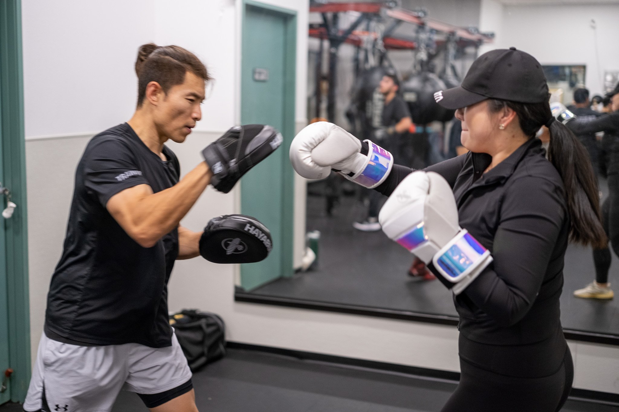 A man and woman practicing boxing in a gym, with the man throwing a punch and the woman sparring with gloves ready, surrounded by gym equipment and others in the background.