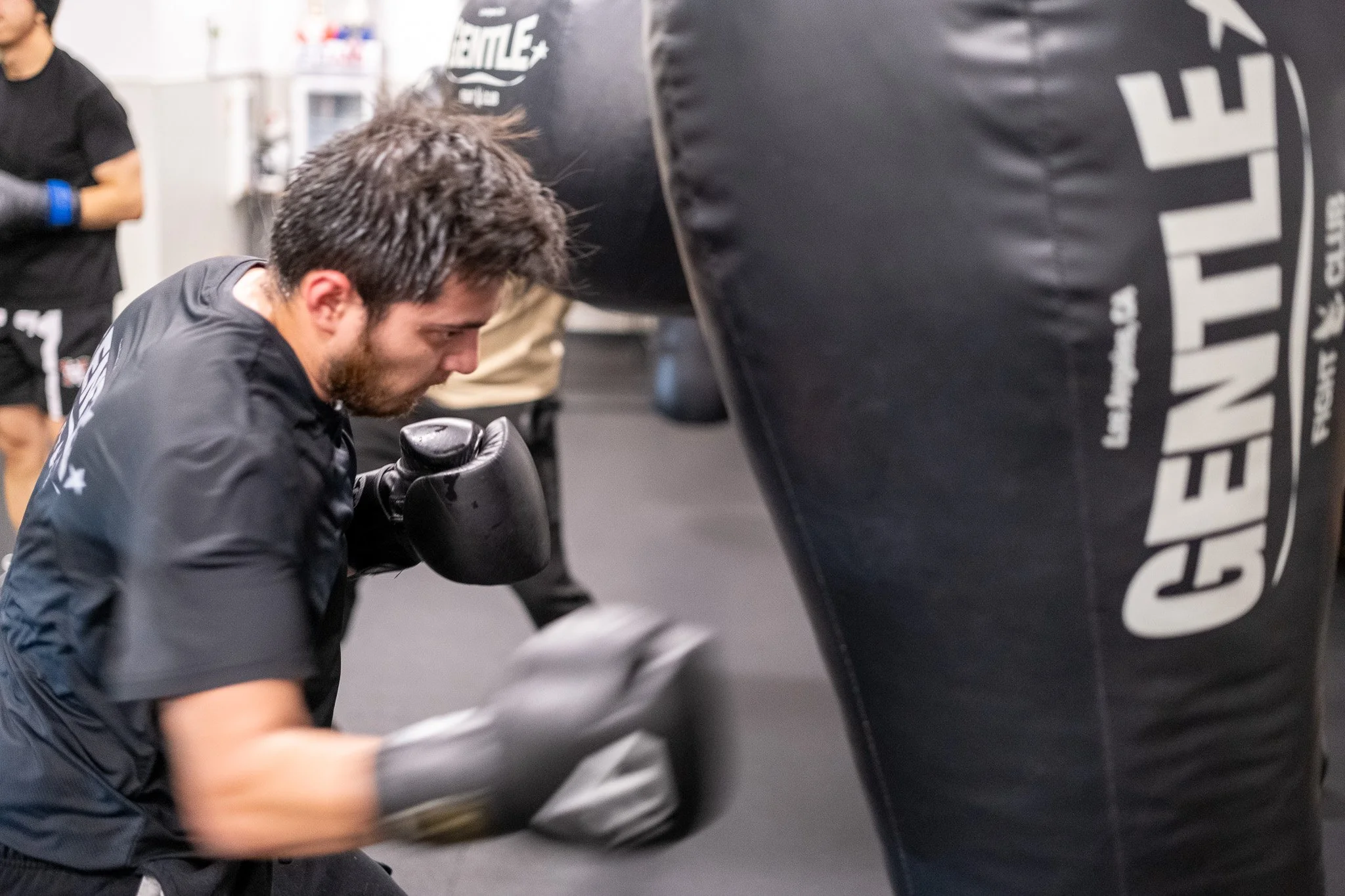 A man wearing black gloves and a black shirt is practicing boxing on a black punching bag in a gym.