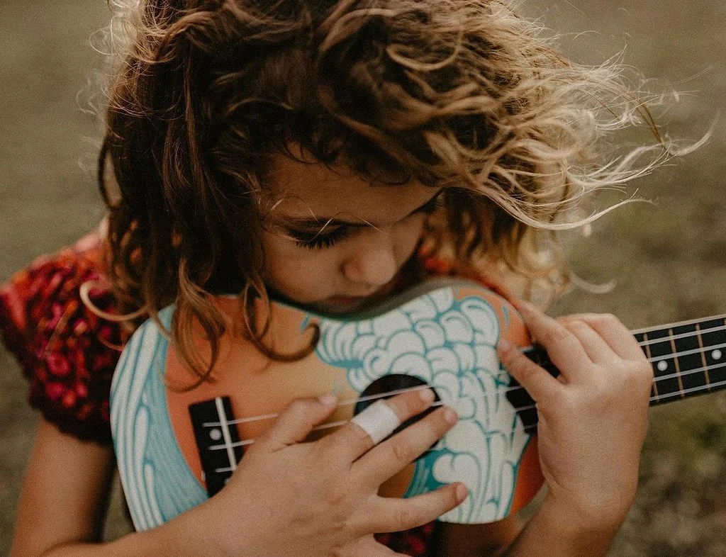 A young girl with curly hair playing a small colorful guitar outdoors.
