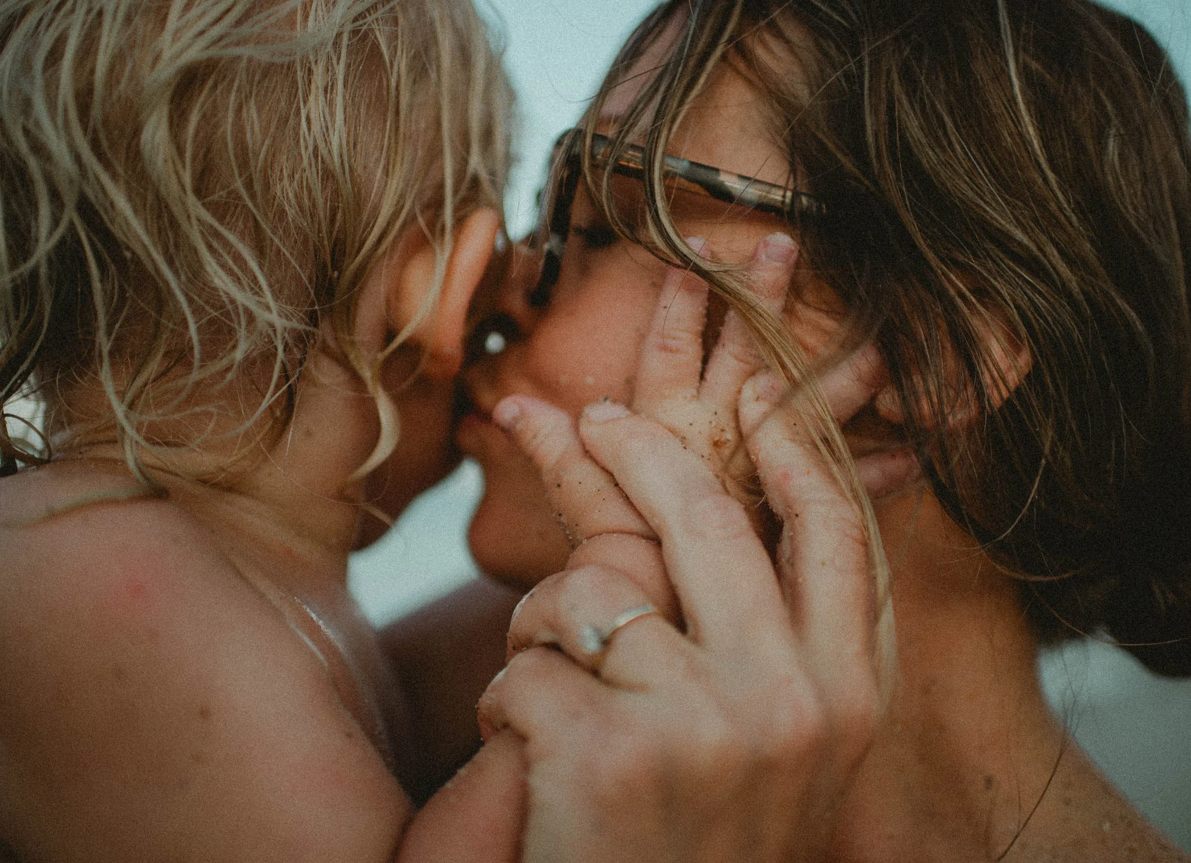 Two women are close together, one woman with curly hair has her hands covering part of her face, while the woman with straight hair and glasses is touching her face. They appear to be intimate and emotional.