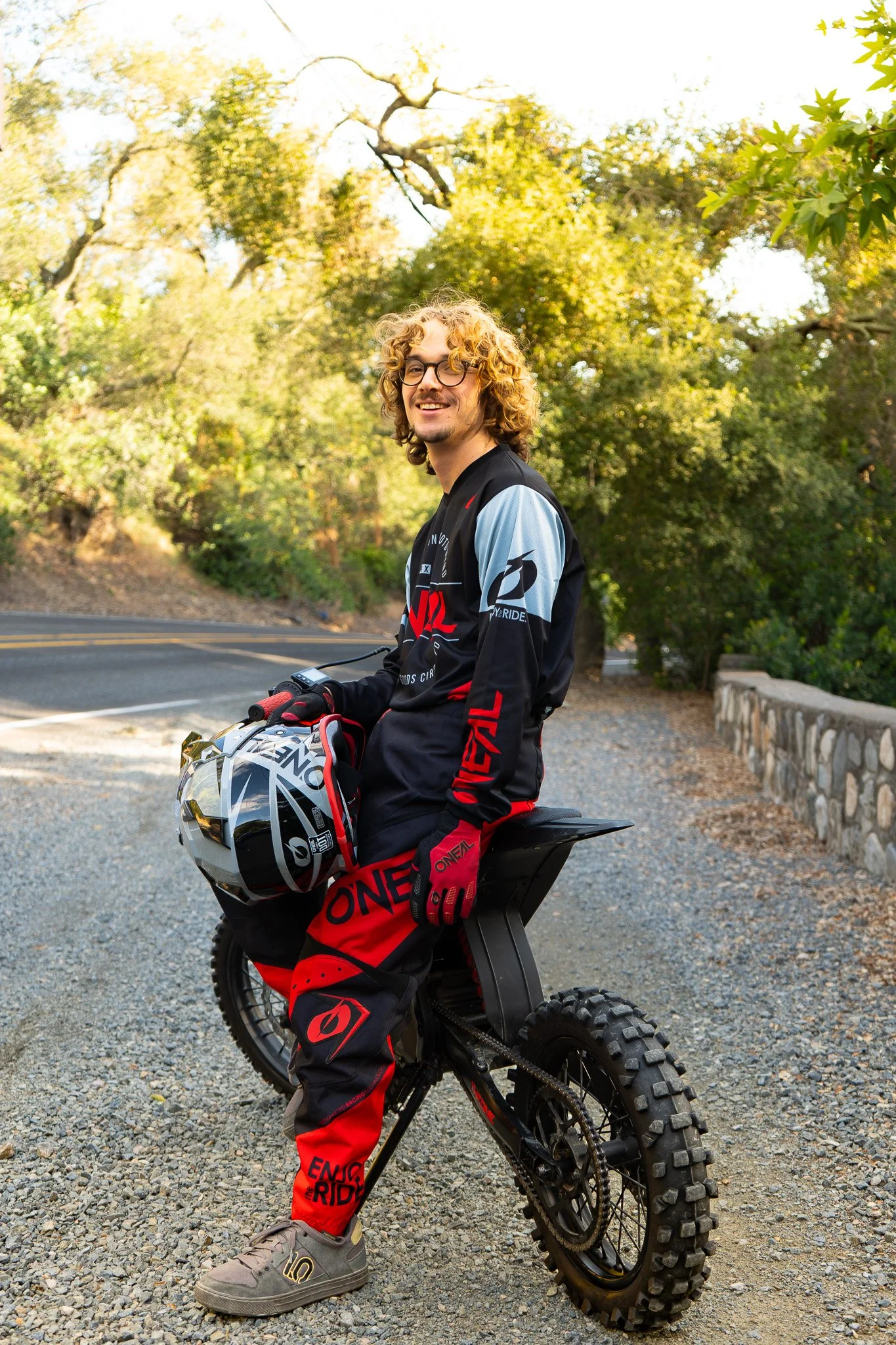 A young man with curly blond hair wearing glasses and a black and red motocross outfit is sitting on a dirt bike holding a helmet. He is outdoors with trees and a paved road in the background.