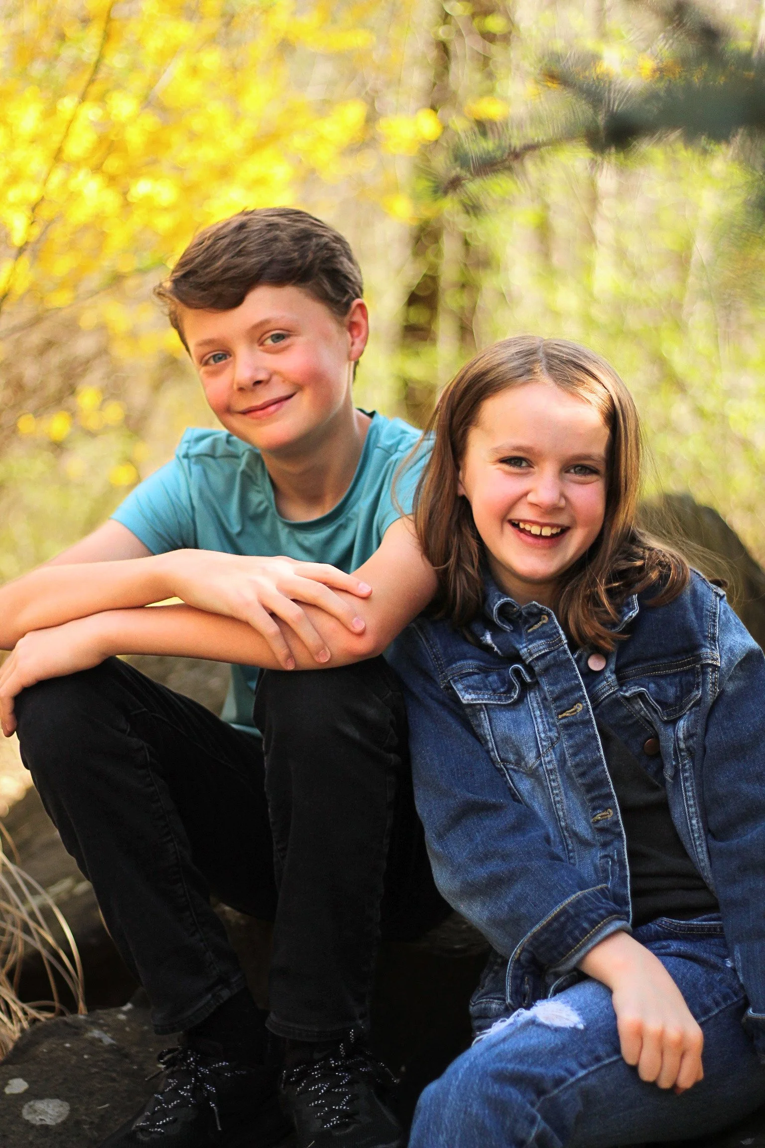 A young boy and girl sitting outdoors among trees with yellow leaves, smiling at the camera.