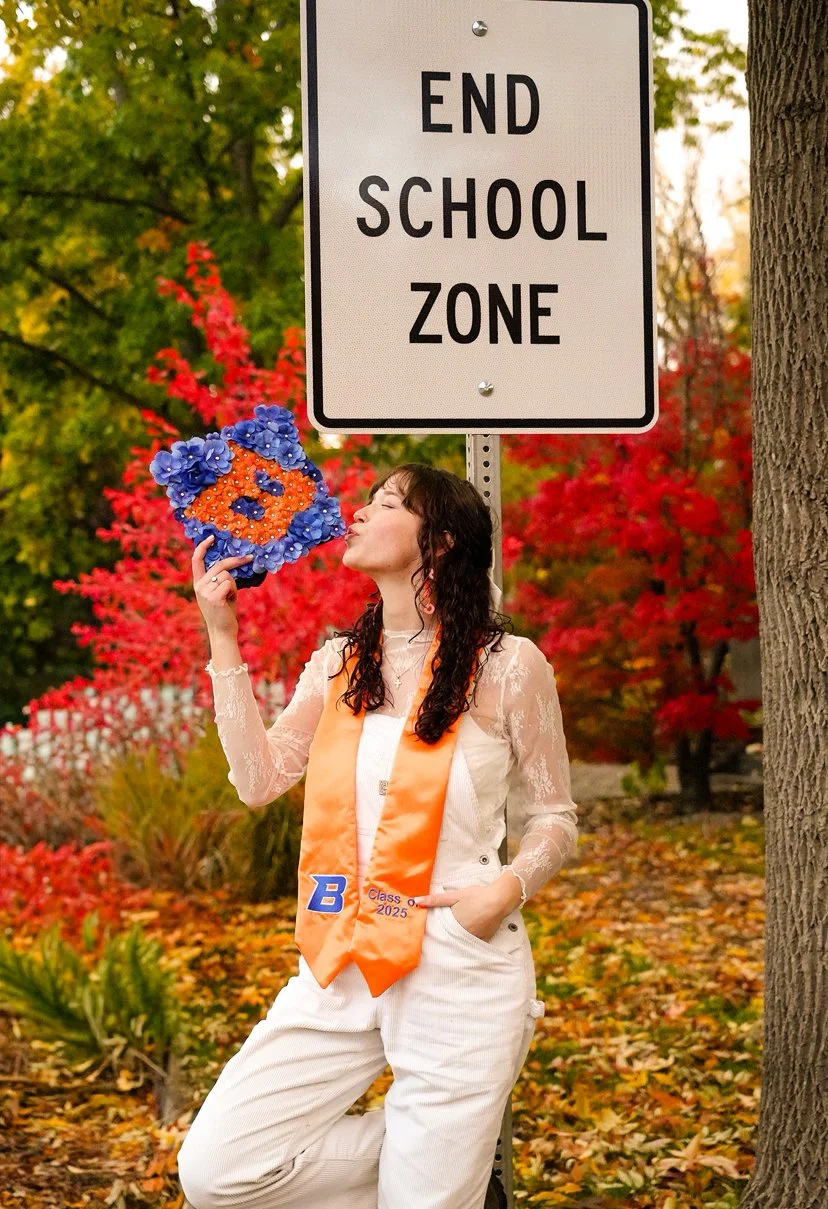 A young woman blowing a kiss holding a decorated flower bouquet in an outdoor setting with fall foliage. She is wearing a white outfit with an orange sash that says "Class of 2025" and a blue letter "B".