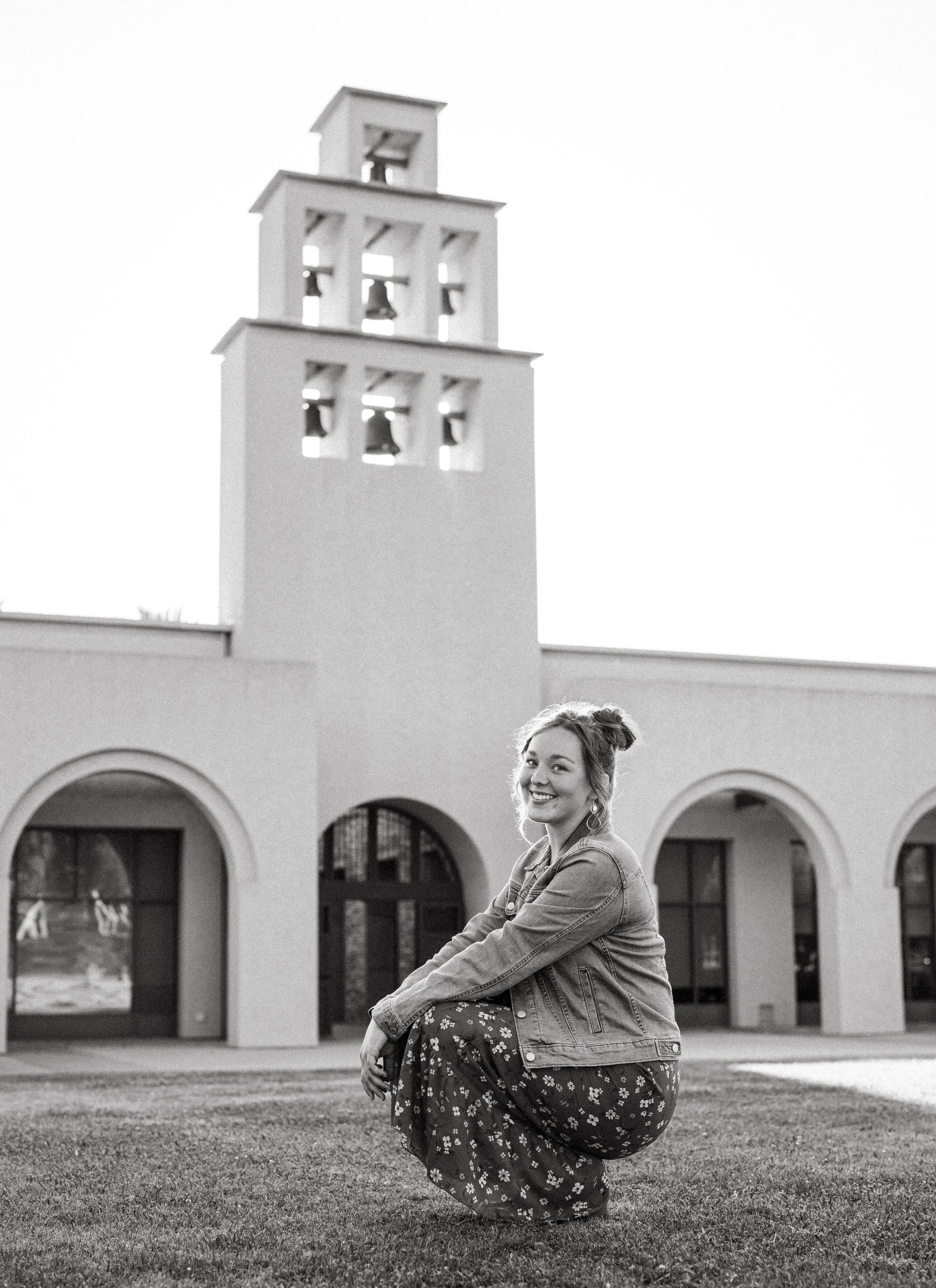 A smiling young woman crouching on grass in front of a building with a tower and arches, wearing a denim jacket and a long floral skirt.