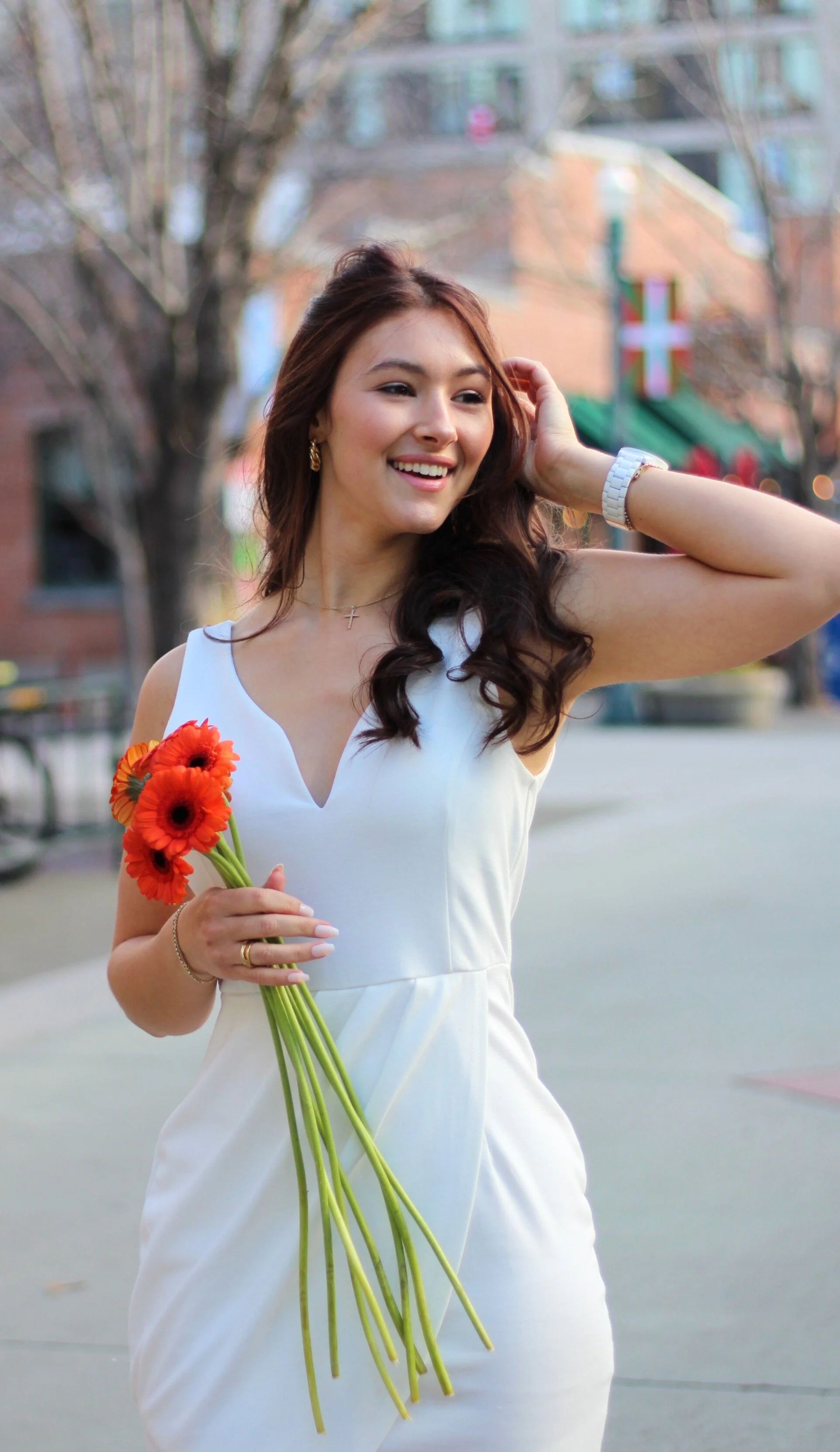 A young woman in a white dress holding a bouquet of orange flowers, smiling outdoors with blurred trees and buildings in the background.