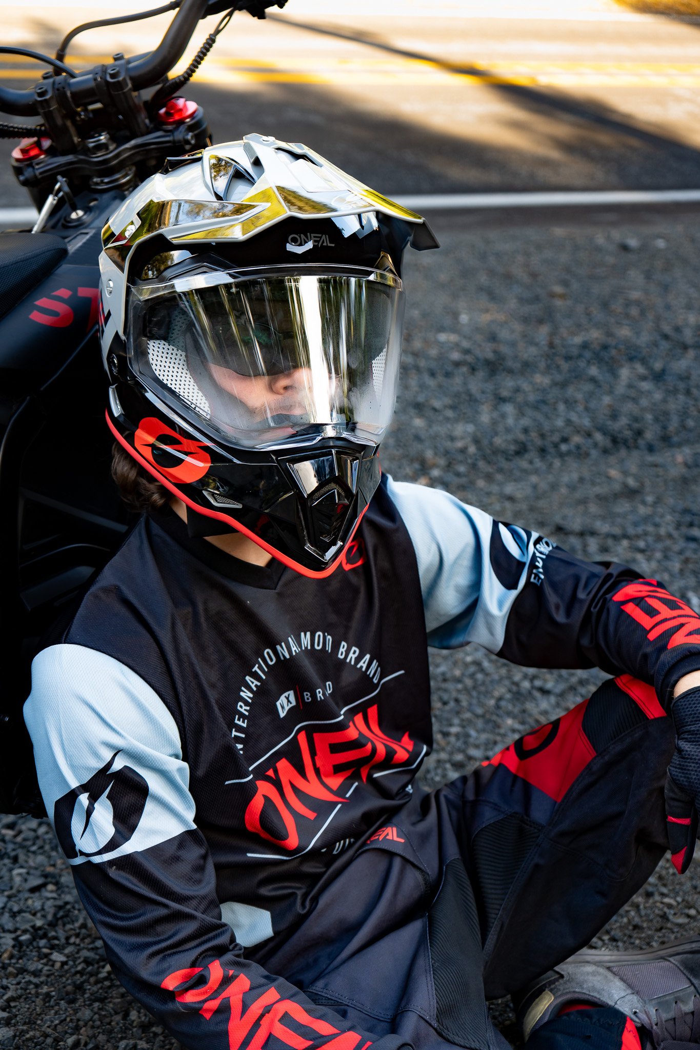 Close-up of a person wearing a black, red, and white motocross helmet and riding gear, sitting on a gravel surface near a road.