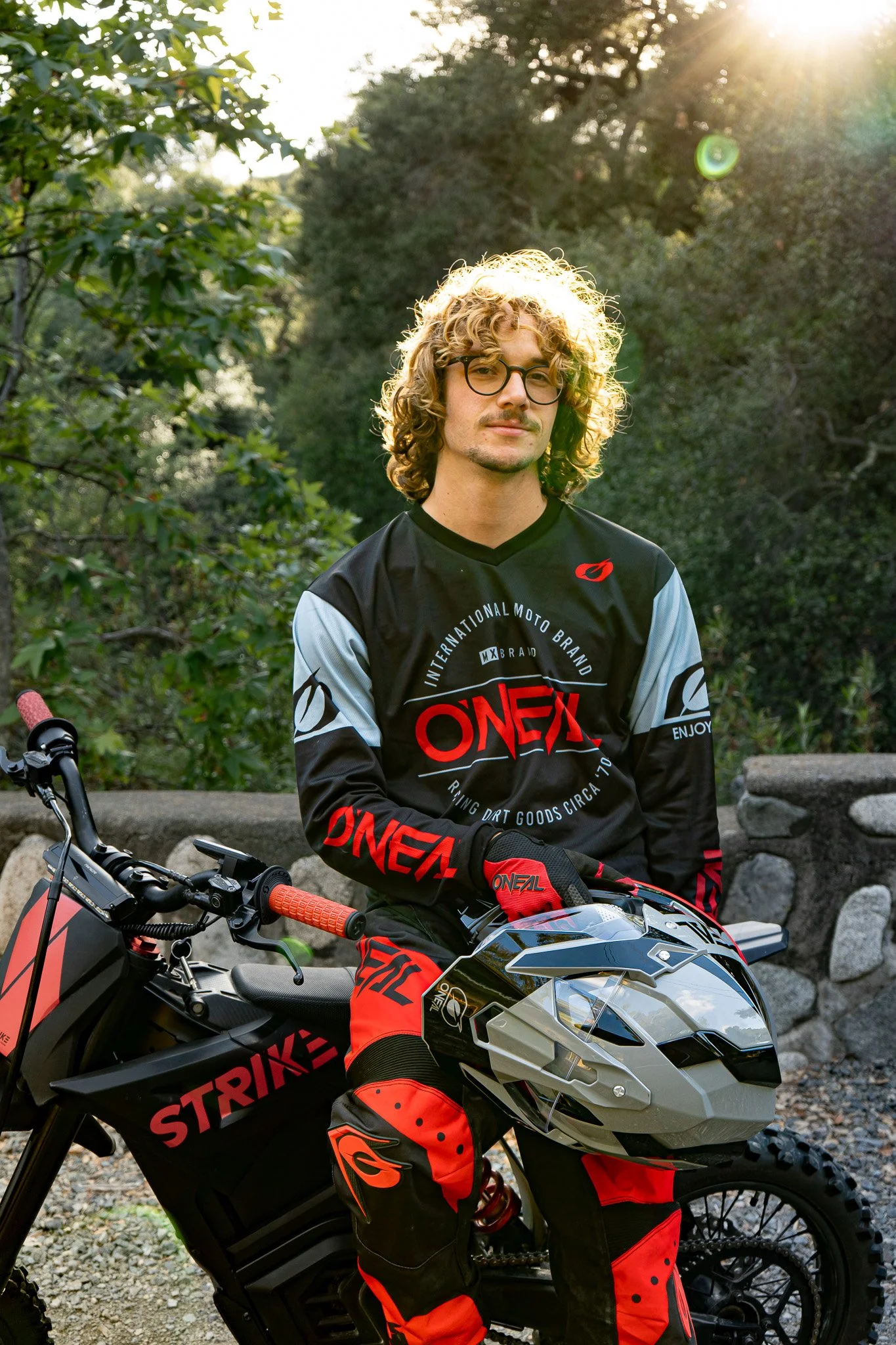 A young man with curly blonde hair and glasses sitting on a black and red dirt bike outdoors in a park-like setting with trees and sunlight.