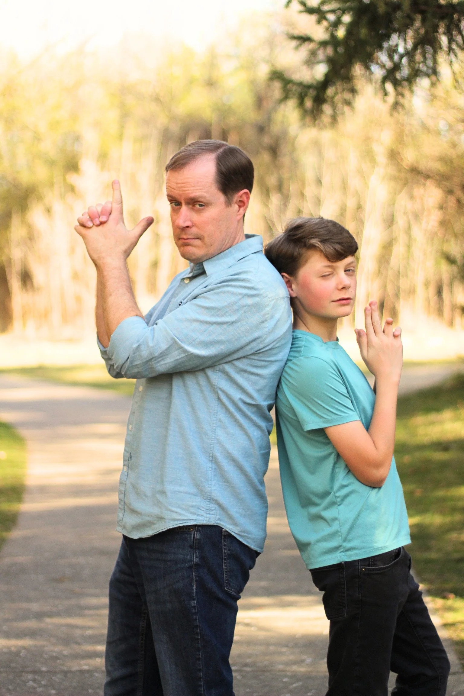 A man and a boy standing back-to-back outdoors in a park, both with arms crossed and hands in gestures, with a blurred background of trees and sunlight.
