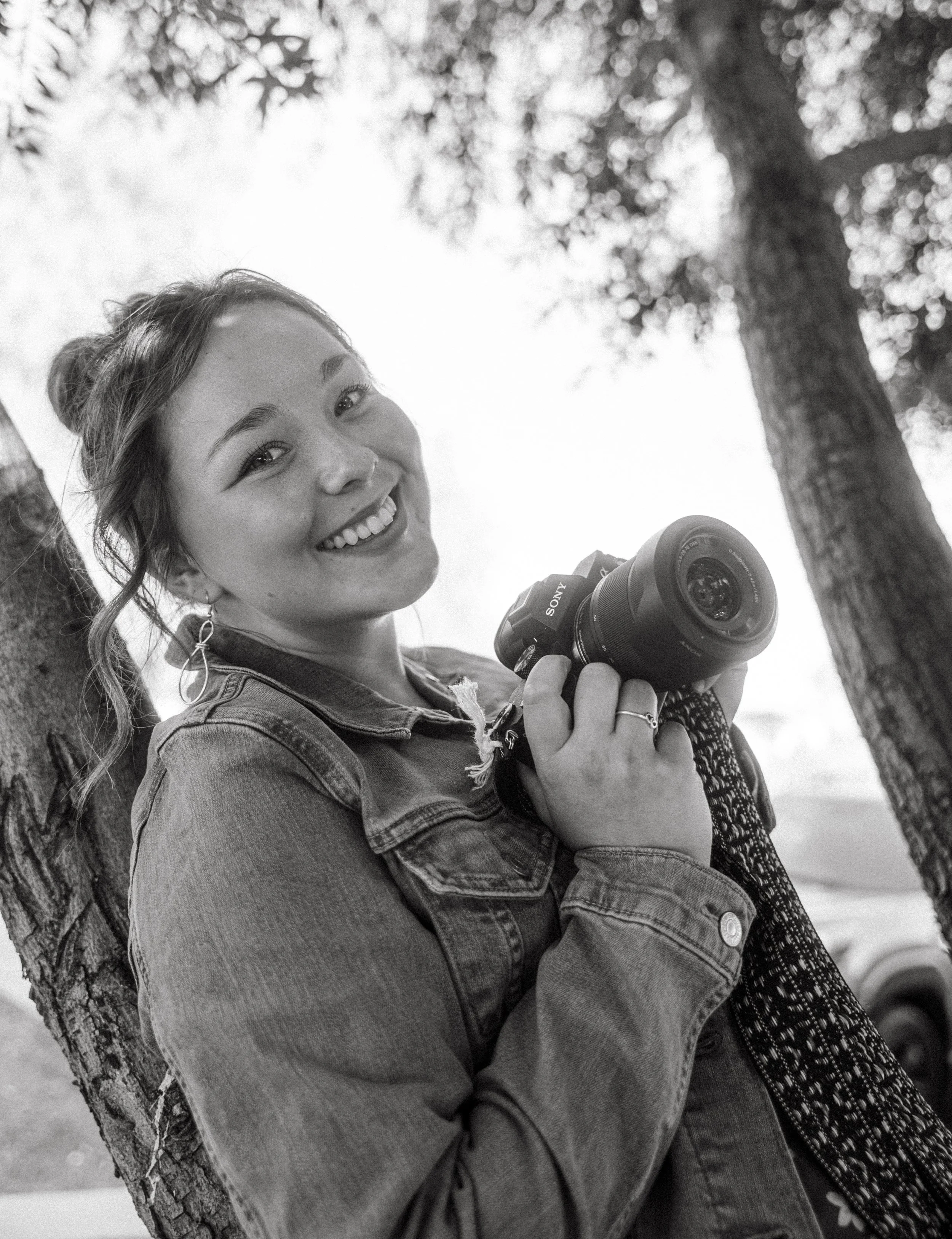 A woman with light skin, smiling, holding a camera, wearing earrings and a denim jacket, standing outdoors near a tree with a blurred background.