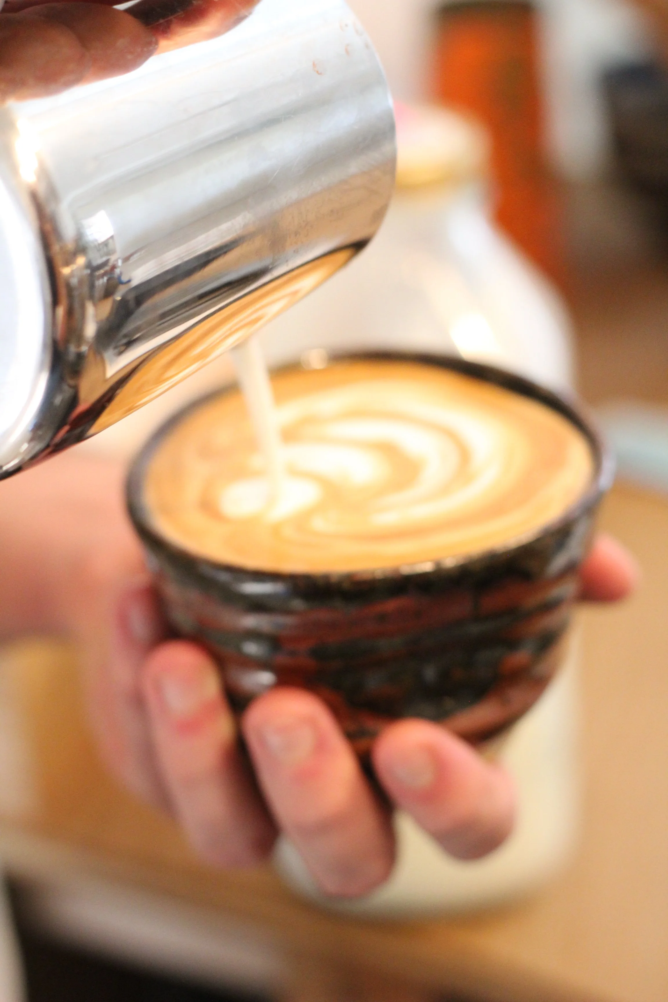 Close-up of a hand holding a small cup of coffee with latte art design on top, with a can being poured into the cup.