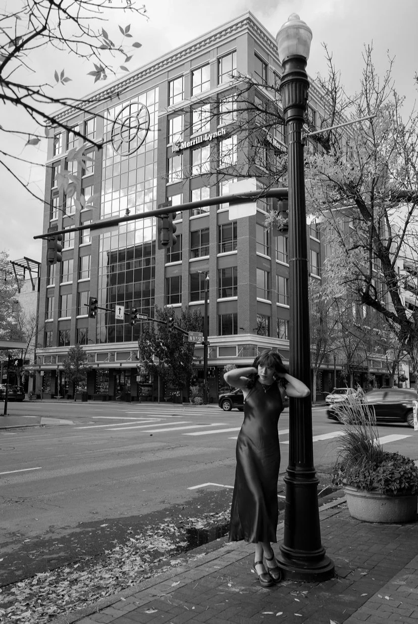 A woman in a long dress stands on a city sidewalk, leaning against a streetlamp, with a large office building in the background. The scene is captured in black and white.