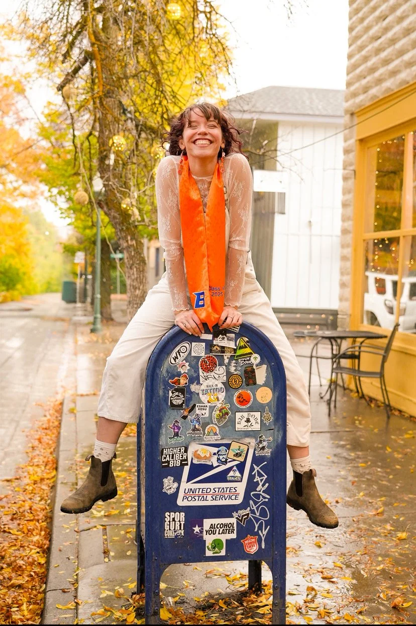 A woman with short brown hair, wearing a sheer lace long-sleeve top, cream-colored pants, and brown boots, is sitting on a blue USPS mailbox on a city sidewalk with fallen autumn leaves. She is smiling broadly and wearing an orange class of 2025 sash. The mailbox is decorated with various stickers.