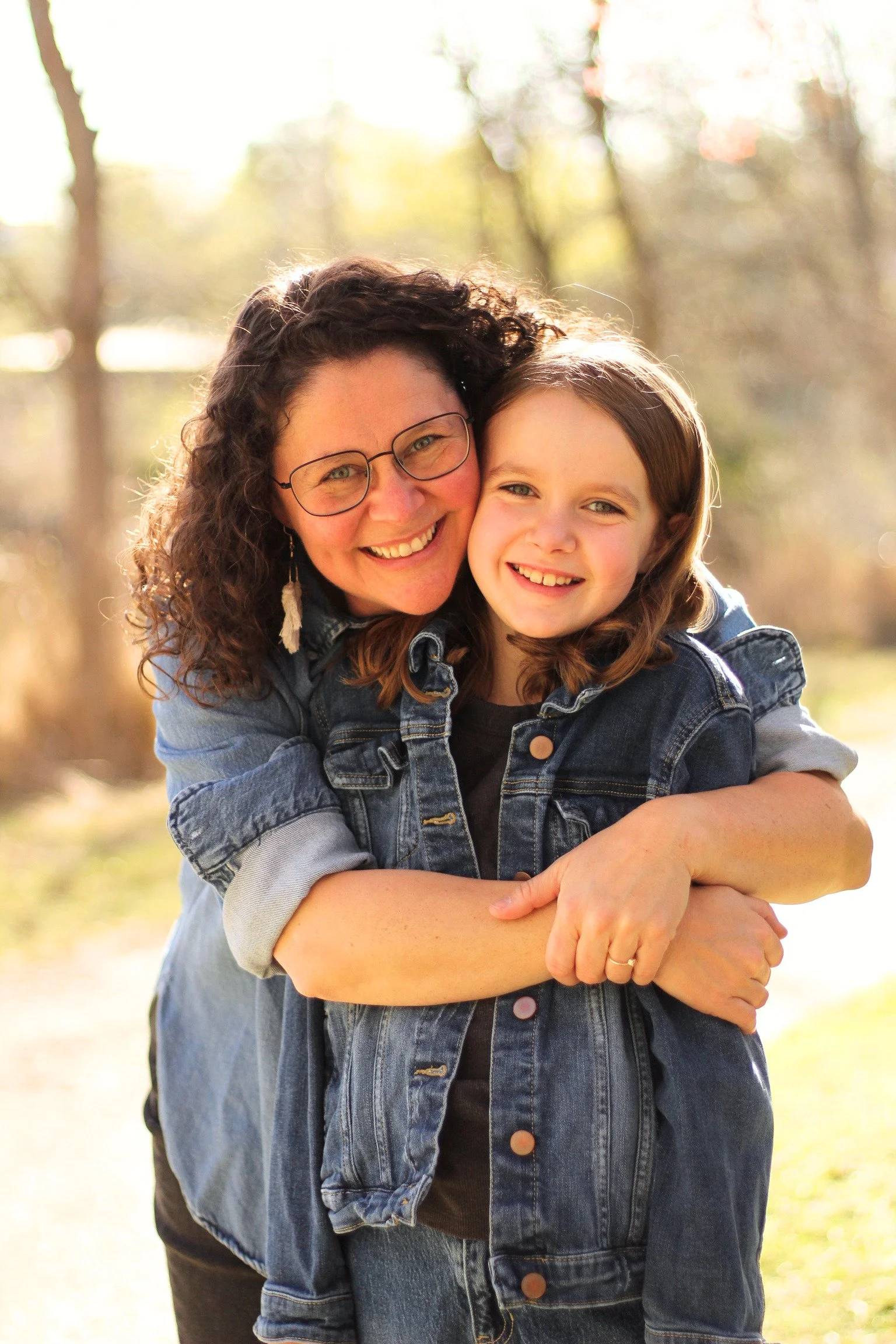 A woman with curly brown hair, wearing glasses and a denim jacket, hugging a young girl with brown hair, also wearing a denim jacket, outdoors with blurred trees and warm sunlight in the background.