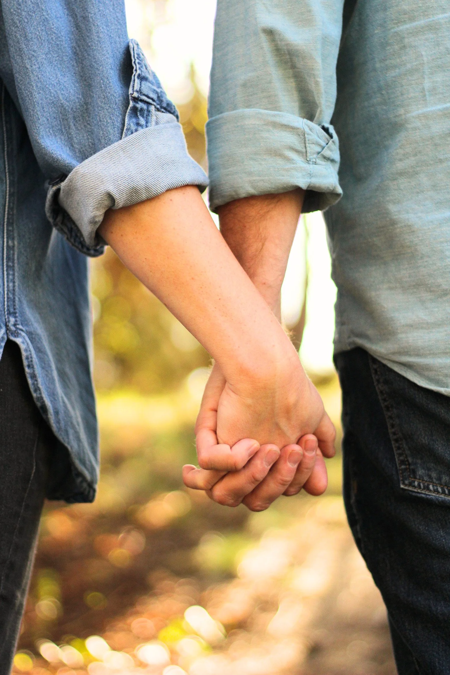 Two people holding hands, one wearing a denim jacket and the other a gray shirt, outdoors with blurred background.