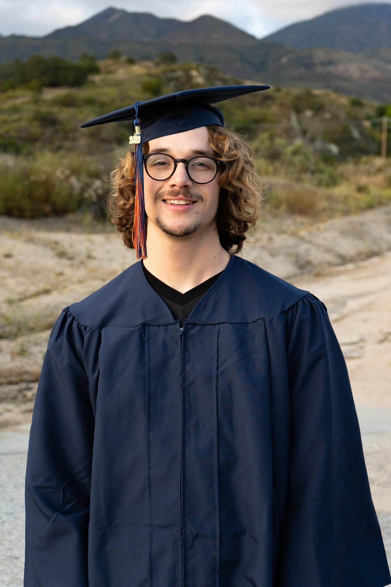 A young man is smiling and wearing a graduation cap and gown outdoors, with mountains and cloudy sky in the background.