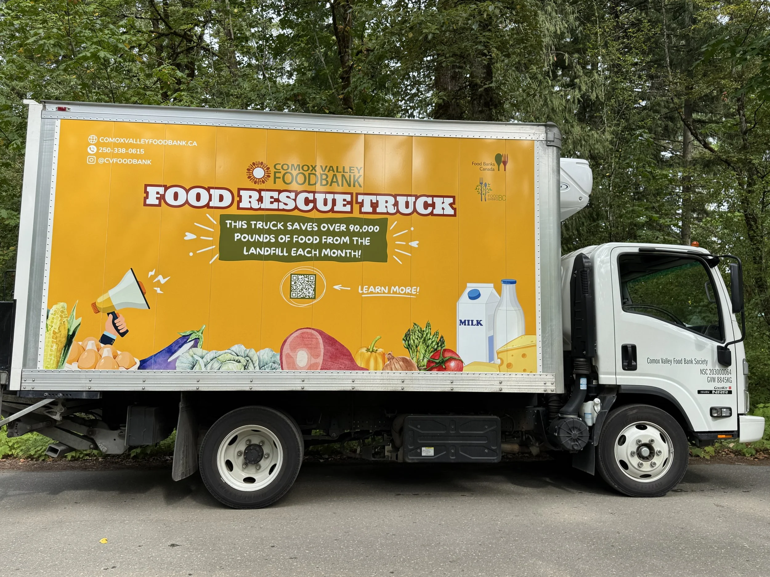 A food rescue truck with a bright orange side panel displaying the logo and information for Comox Valley Food Bank. The panel includes a website, phone number, social media handle, and a QR code. The truck is parked on the street with a background of green trees.