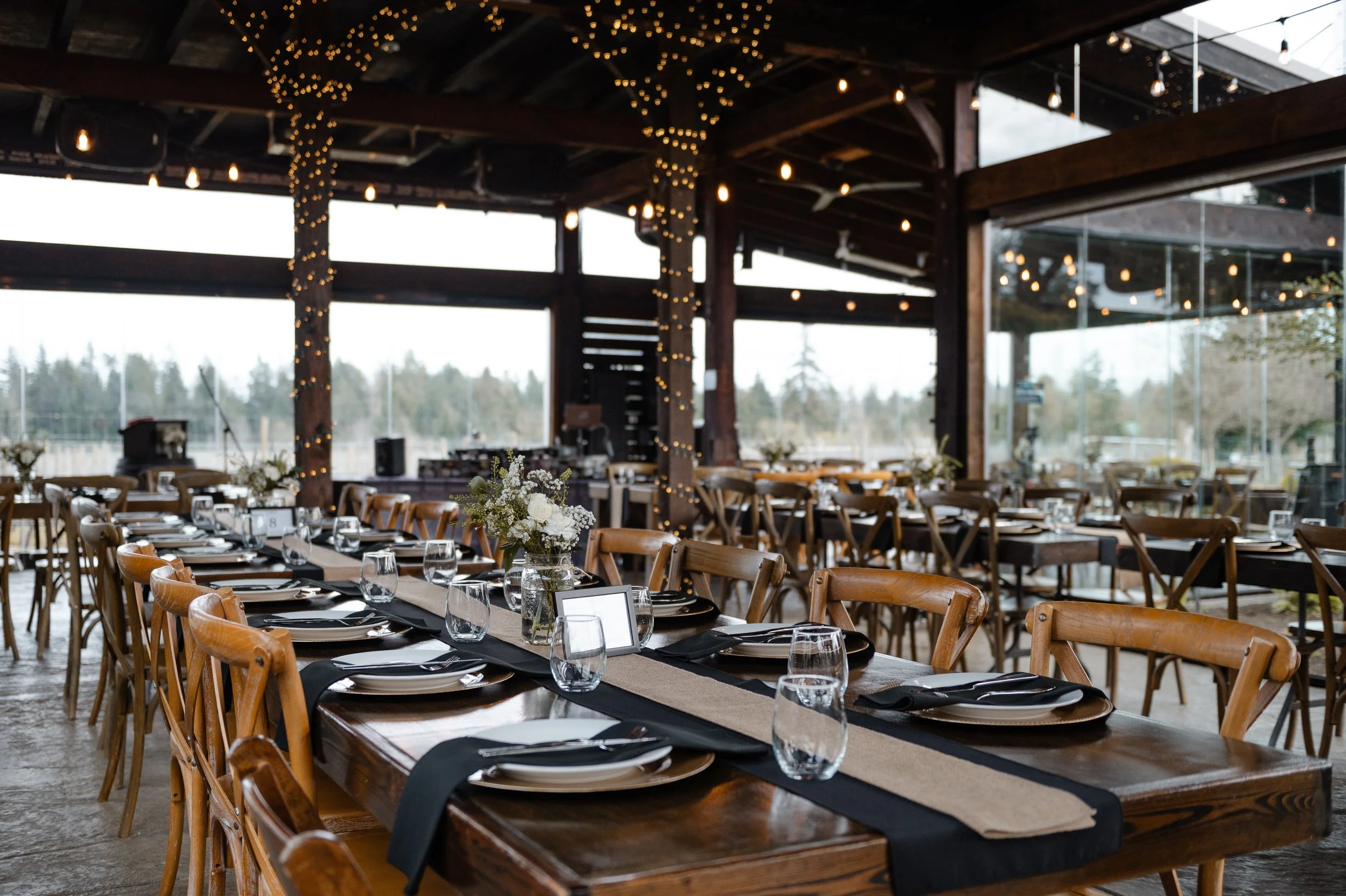 Interior of a rustic event space with round tables set with black table runners, glassware, and floral centerpieces, featuring large windows, wooden beams, string lights, and a view of a landscape outside.