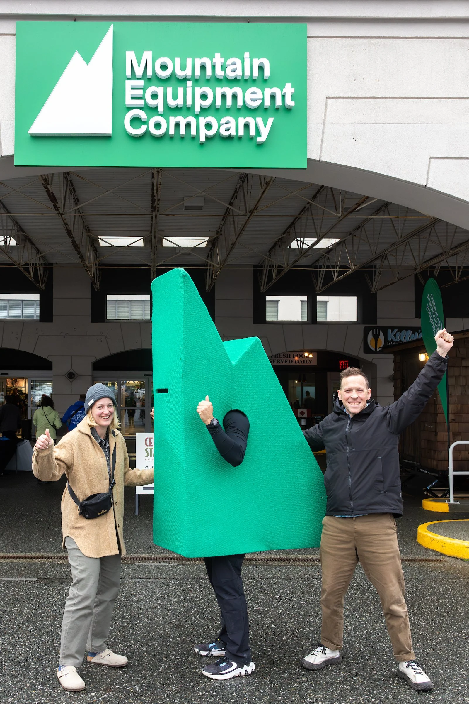 Three smiling people standing in front of a building, holding a large green cutout of a mountain with the logo of Mountain Equipment Company on the sign above.