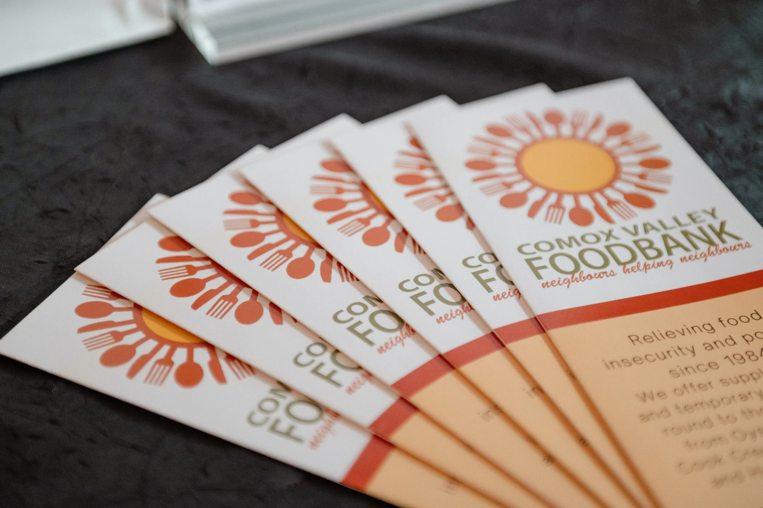 Multiple brochures for Comox Valley Food Bank, featuring a graphic of a sun made from orange utensils on a white background, spread out on a dark surface.
