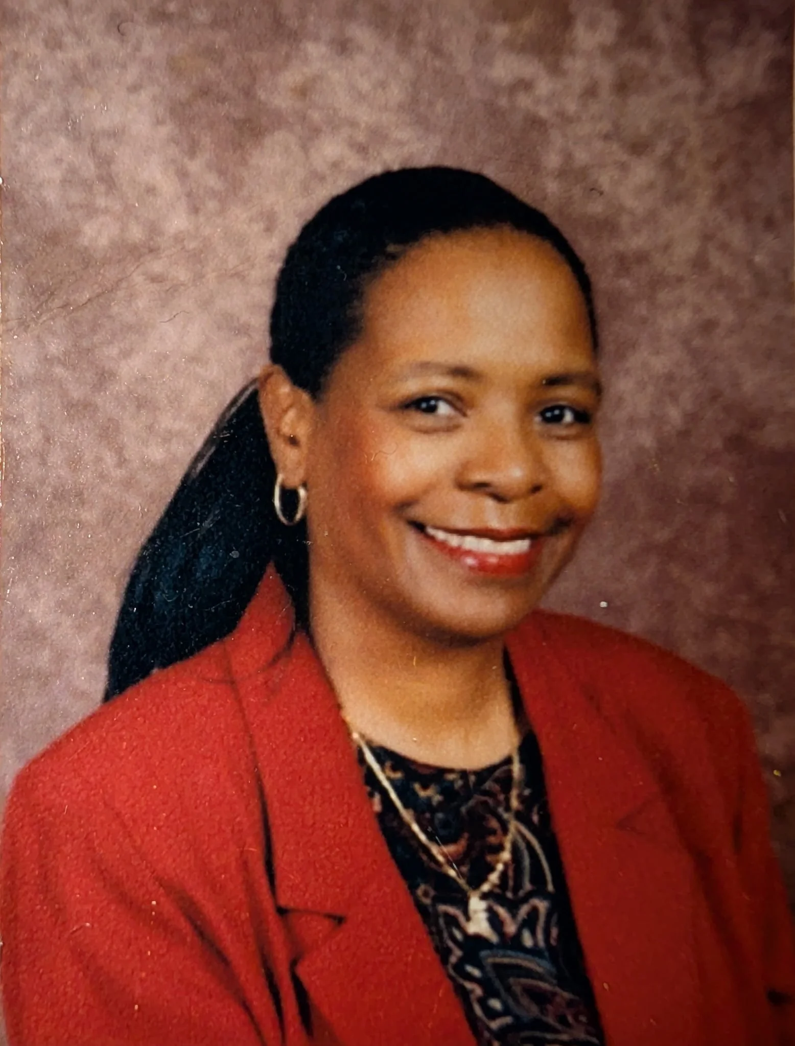 Close-up of a smiling woman with dark hair, wearing a red blazer, earrings, and a patterned blouse, against a textured brown background.