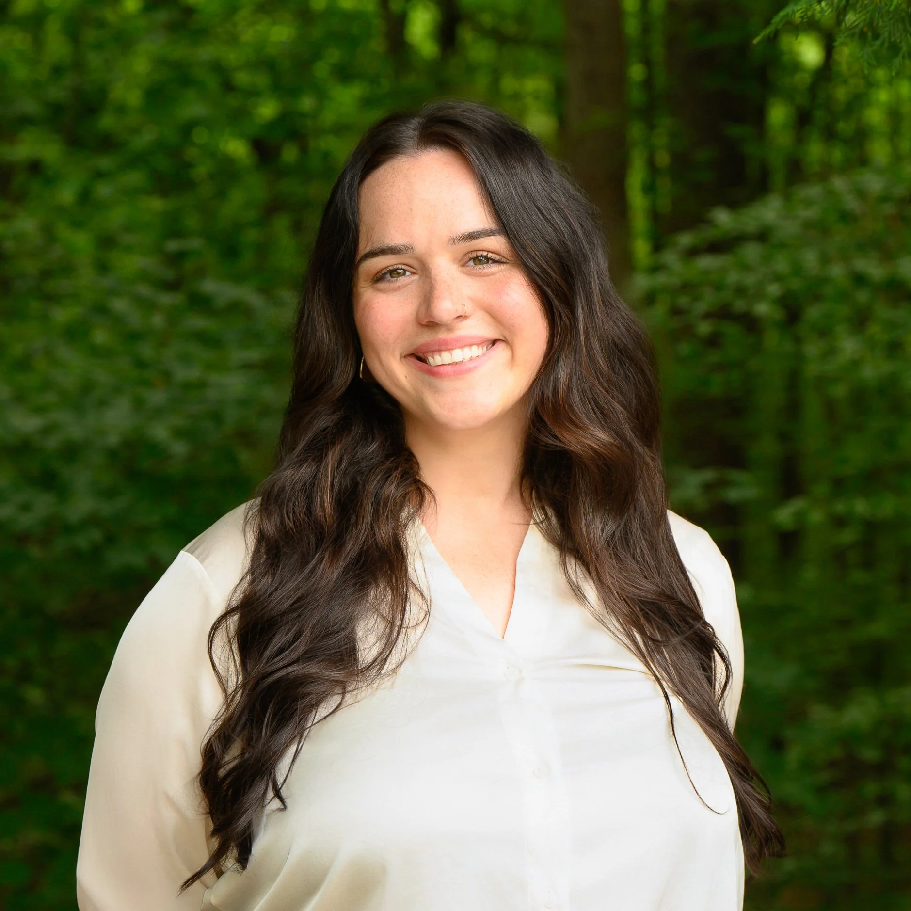 A young woman with long dark hair smiling outdoors in front of a green forest background.
