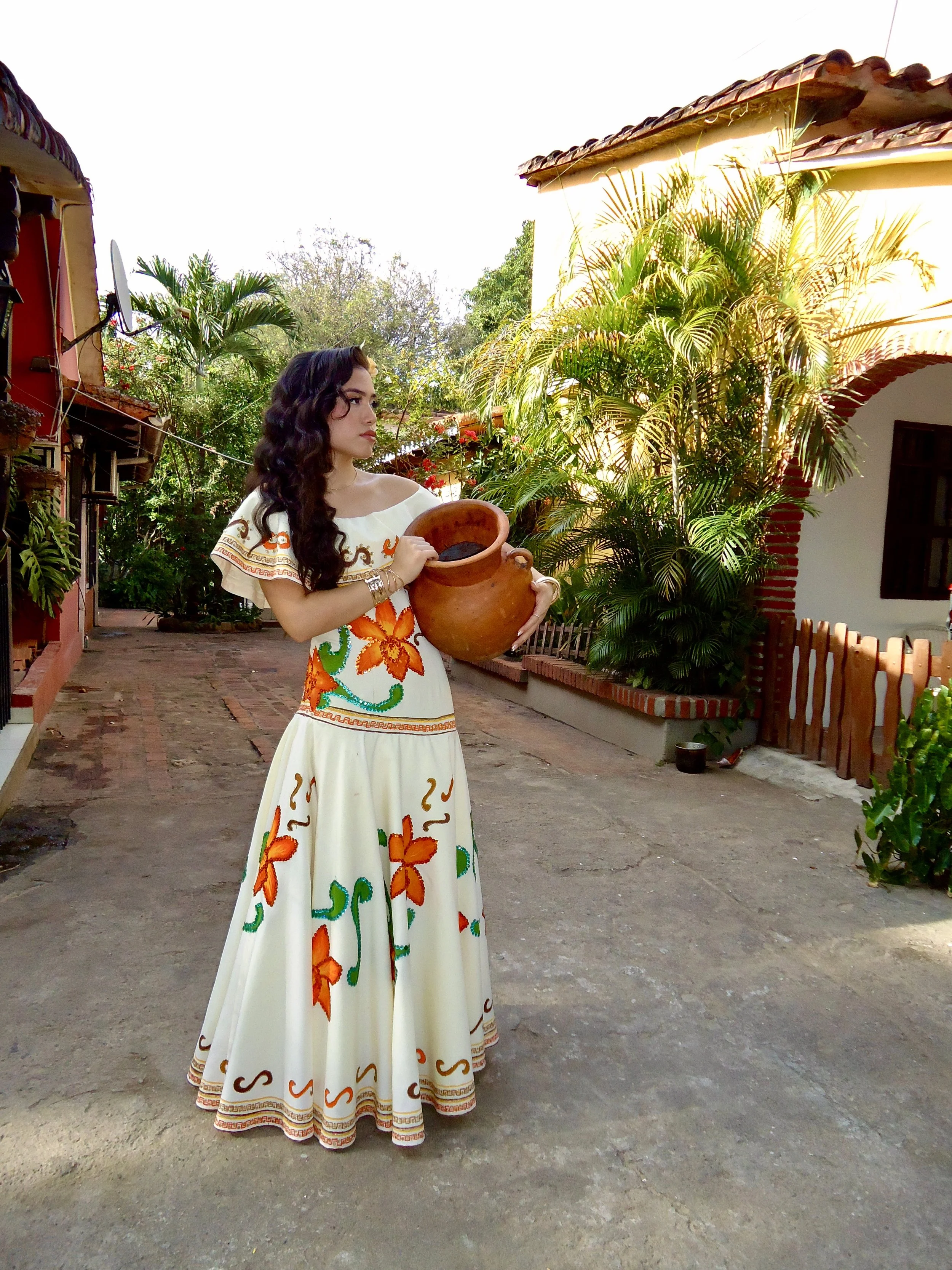 An outdoor photo of a woman wearing a traditional cultural flowy shoulderless dress with painted orange flowers on it while posing for a picture outdoor between two traditional casitas with plants and palm trees in the background