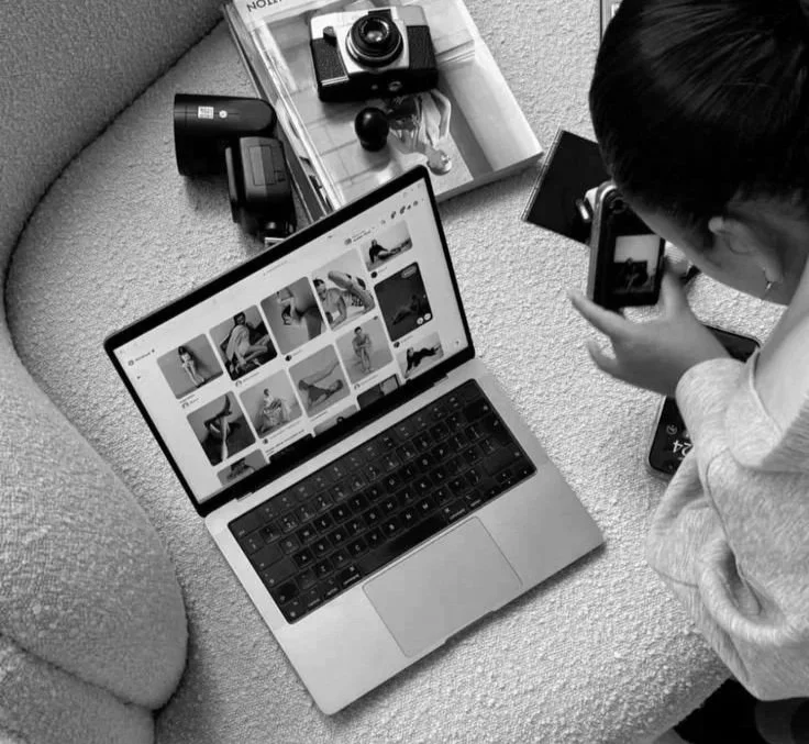 A person taking a photo of their phone with a digital camera, with a laptop displaying a grid of images on a carpeted surface.