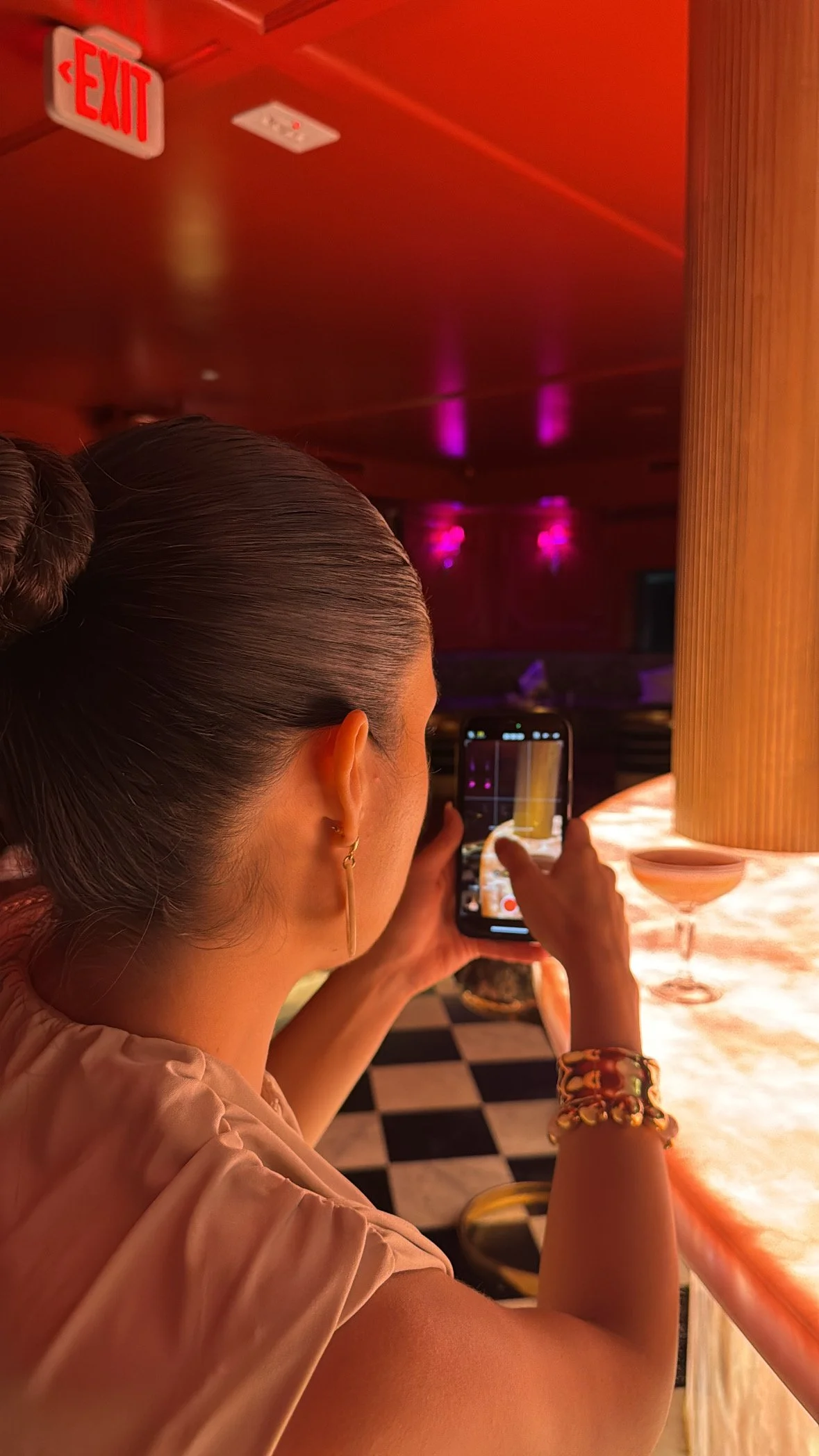 A woman with dark, slicked-back hair in a bun, wearing a beige top, gold earrings, and bracelets, is taking a photo or video with her smartphone at a bar or restaurant. The background features a illuminated bar countertop with a martini glass and a checkered floor, with pink and purple lighting.