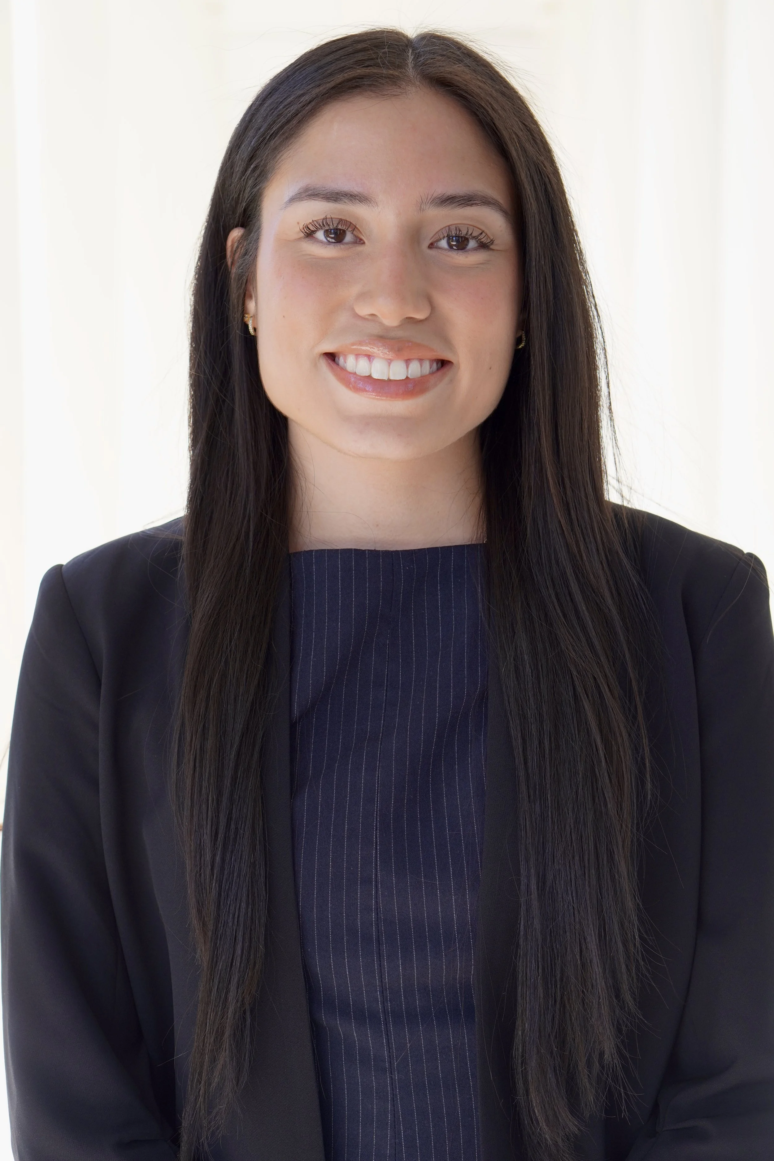 Portrait of a young woman with long dark hair, smiling, wearing a navy blue pinstripe top and black blazer, standing in front of a light-colored background.