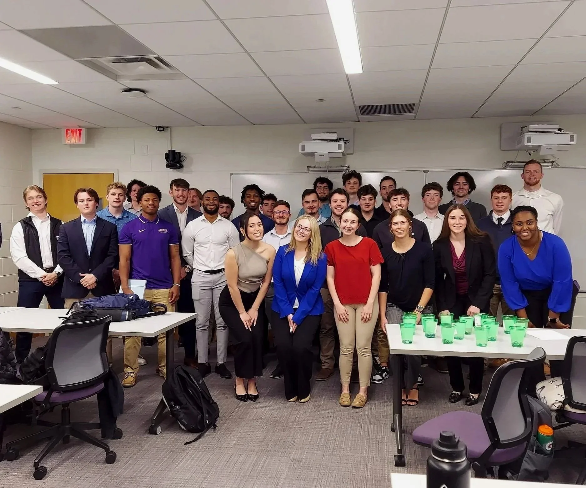 Group of young men and women in a classroom or conference room, standing together and smiling, with tables and chairs around some with backpacks and a table with green cups in the foreground.