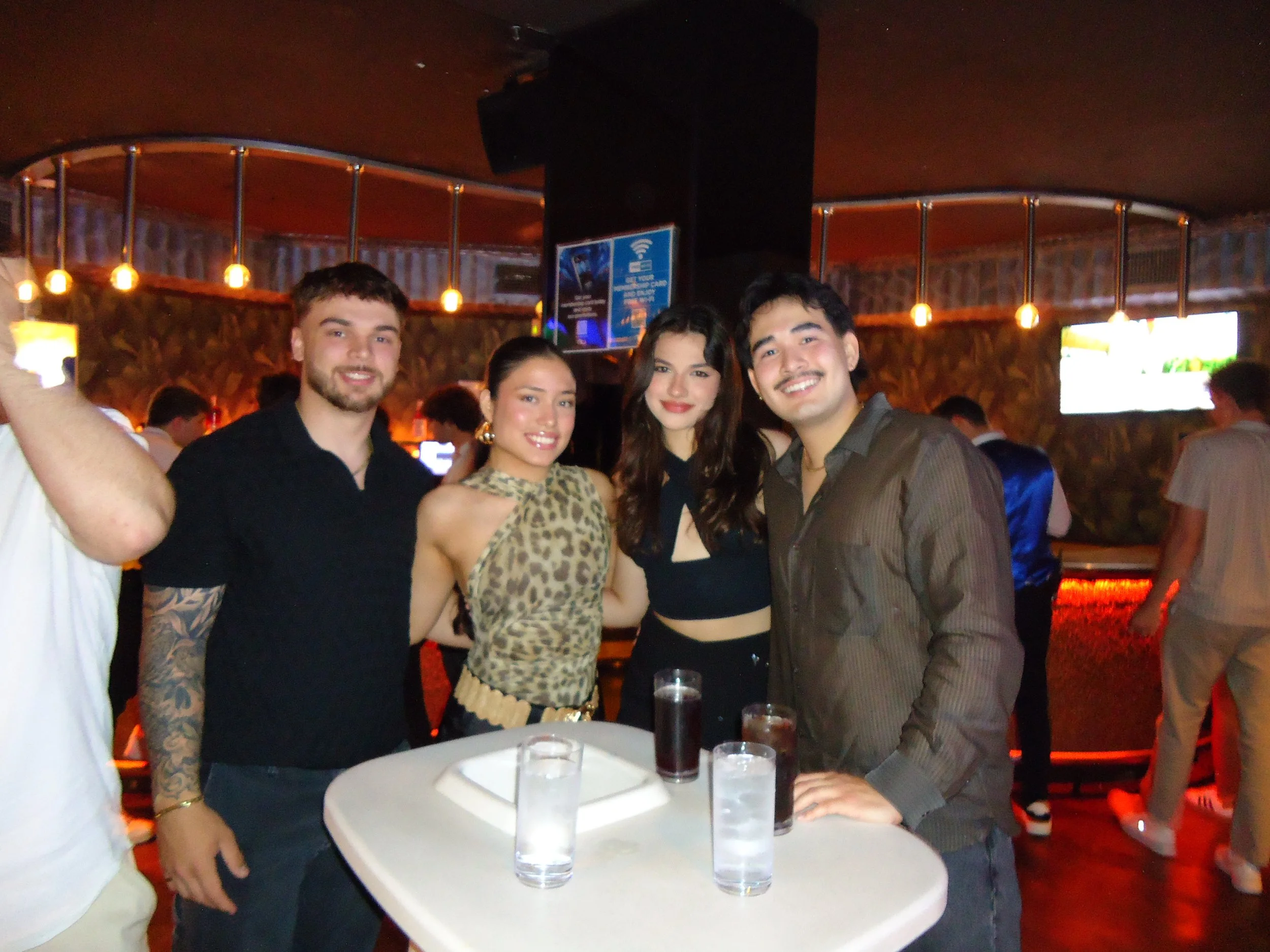 A group of friends, two girls in the middle, two guys on the side of each of them all smiling behind a small white table with drinks on it within a dimmed light bar