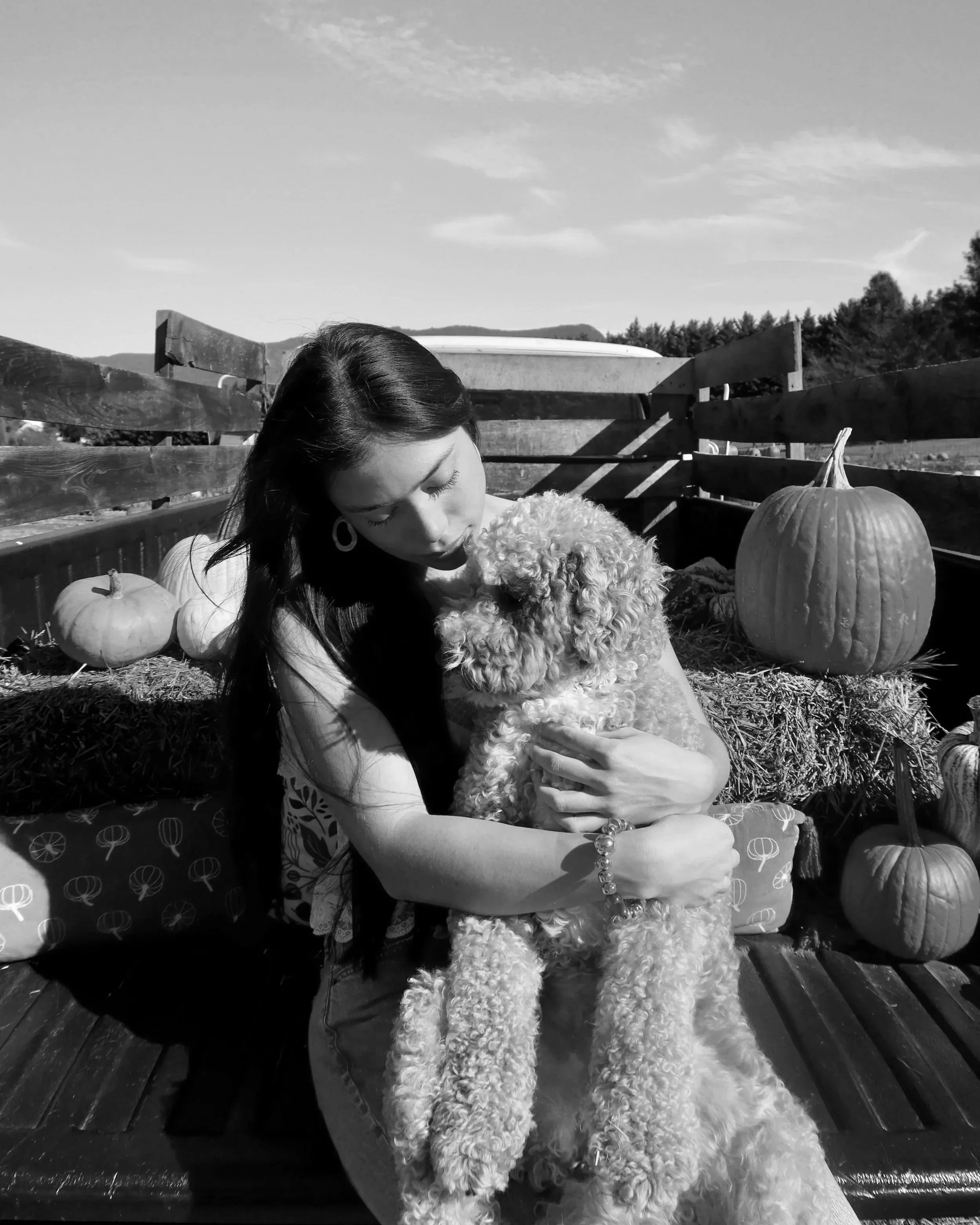 A woman holding a curly-haired dog in an outdoor pumpkin patch setting with pumpkins and hay bales, on a bench, during daytime.