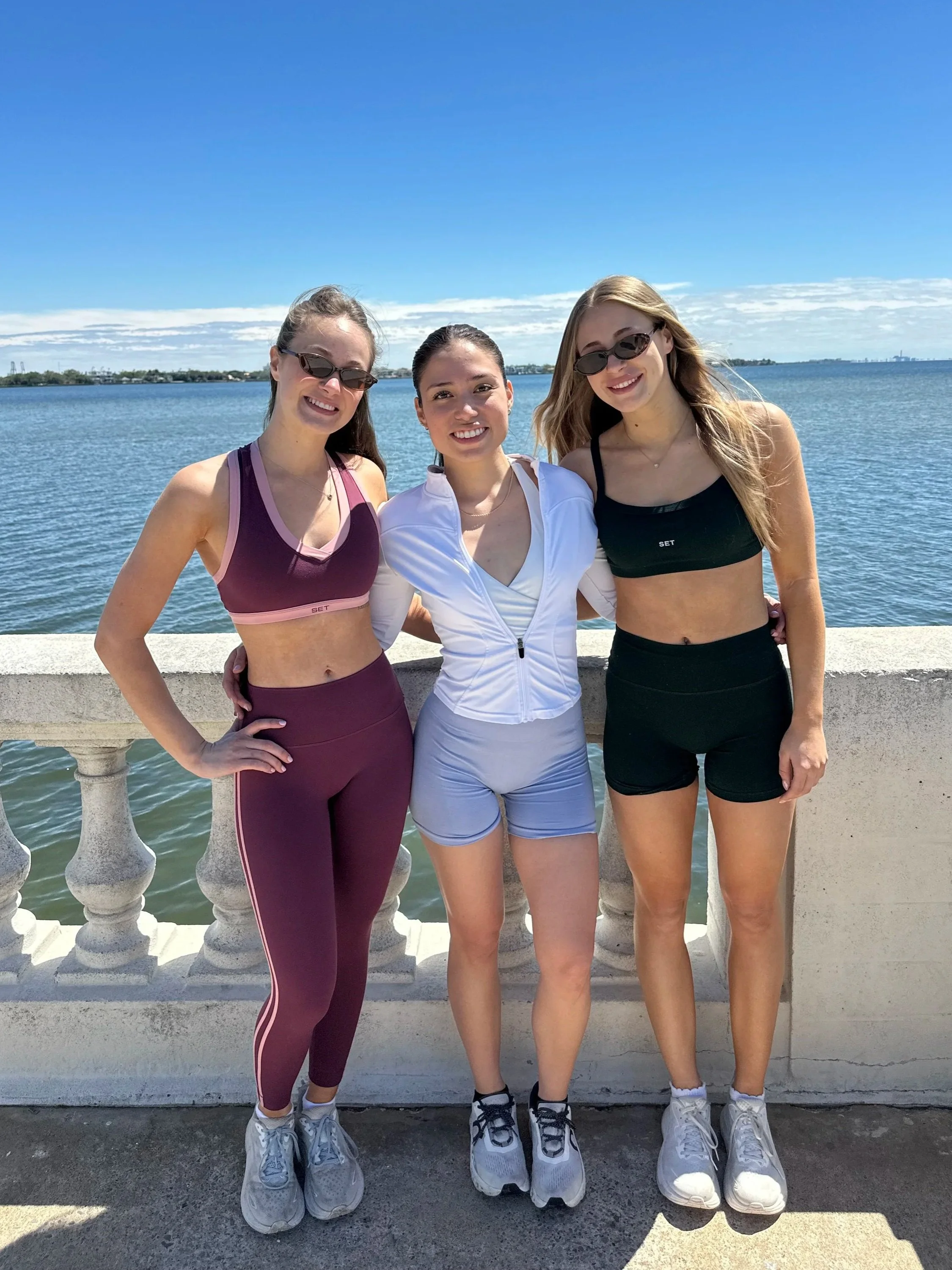 Three girls smiling standing infront of a Bay while wearing their workout sets 