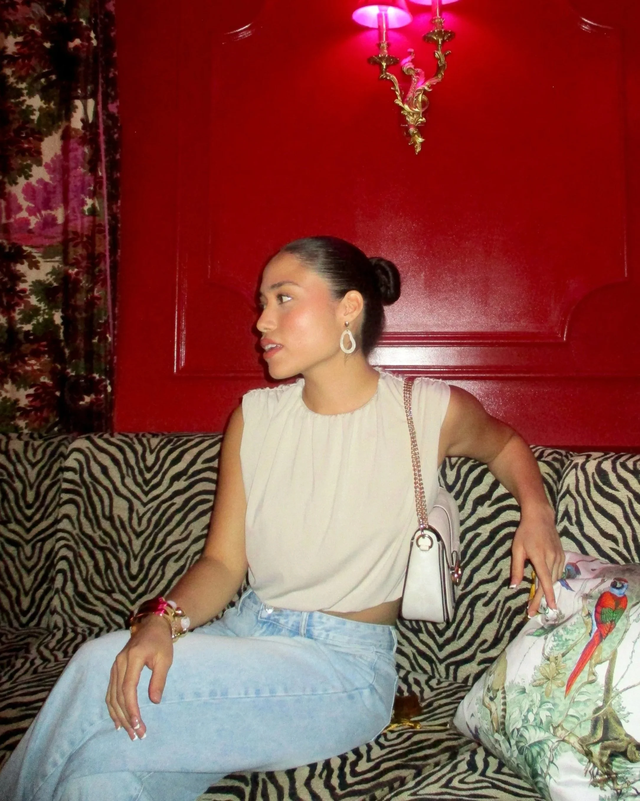 A woman with dark hair tied in a bun, wearing a sleeveless beige top, light blue jeans, and earrings, sitting on a zebra-print sofa with a red wall behind her. She has one arm resting on her lap and the other on a pillow with a colorful parrot design. A chandelier hangs above her, casting pink light.