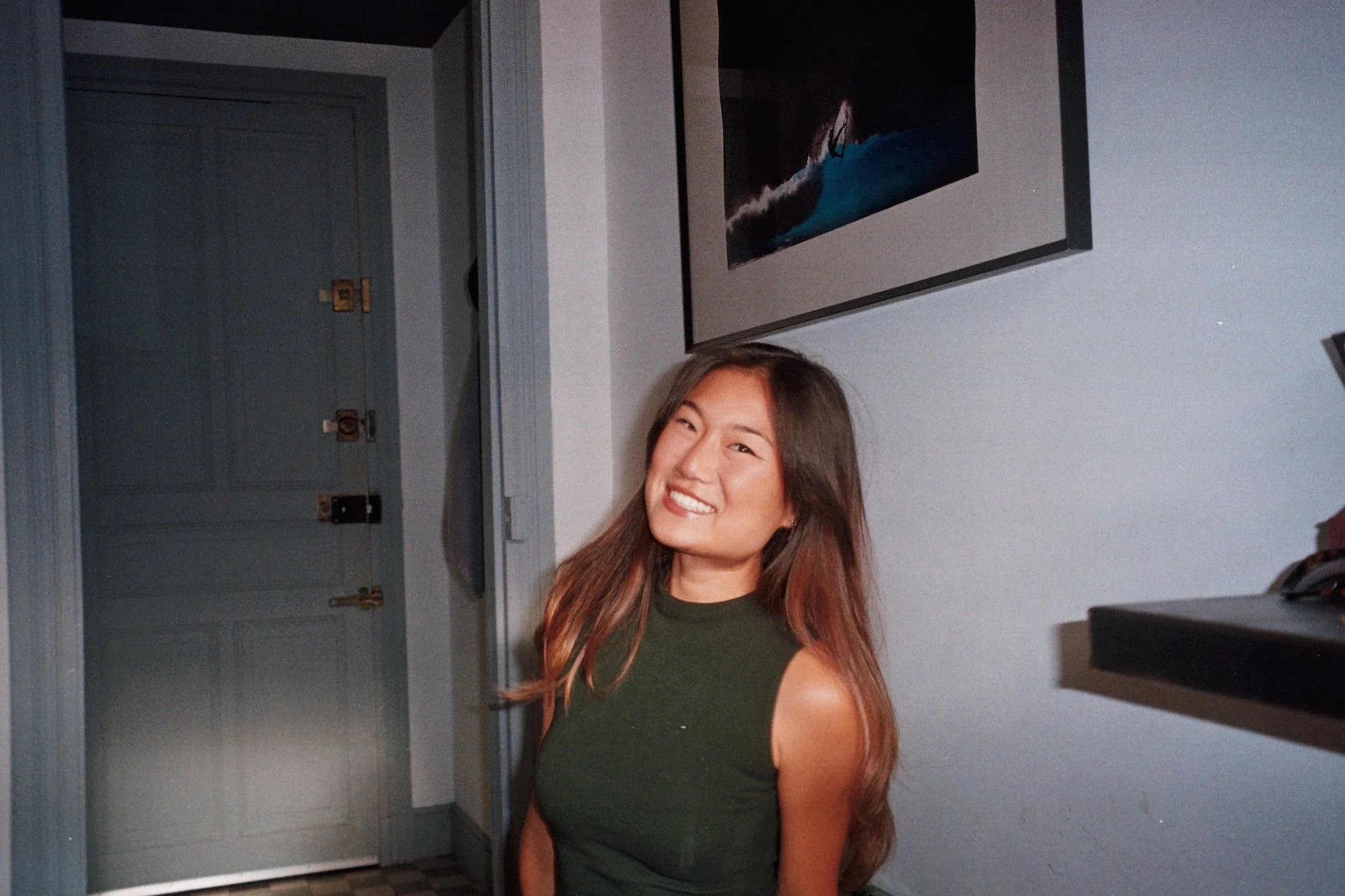 A young woman with long brown hair wearing a sleeveless dark green top, smiling and winking at the camera in a room with a white wall, a painting of a surfer riding a wave, and a closed door.