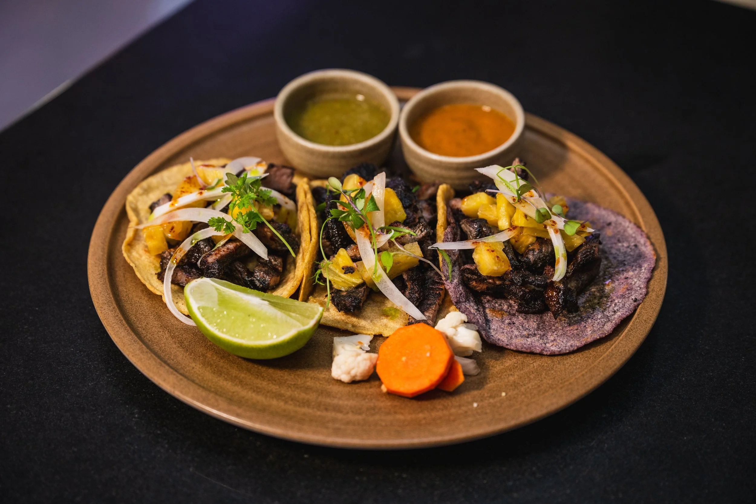 Plate of three street tacos filled with grilled meat, garnished with onions, cilantro, pineapple, and microgreens, with lime wedges, a carrot piece, and two small bowls of salsa on a brown plate.