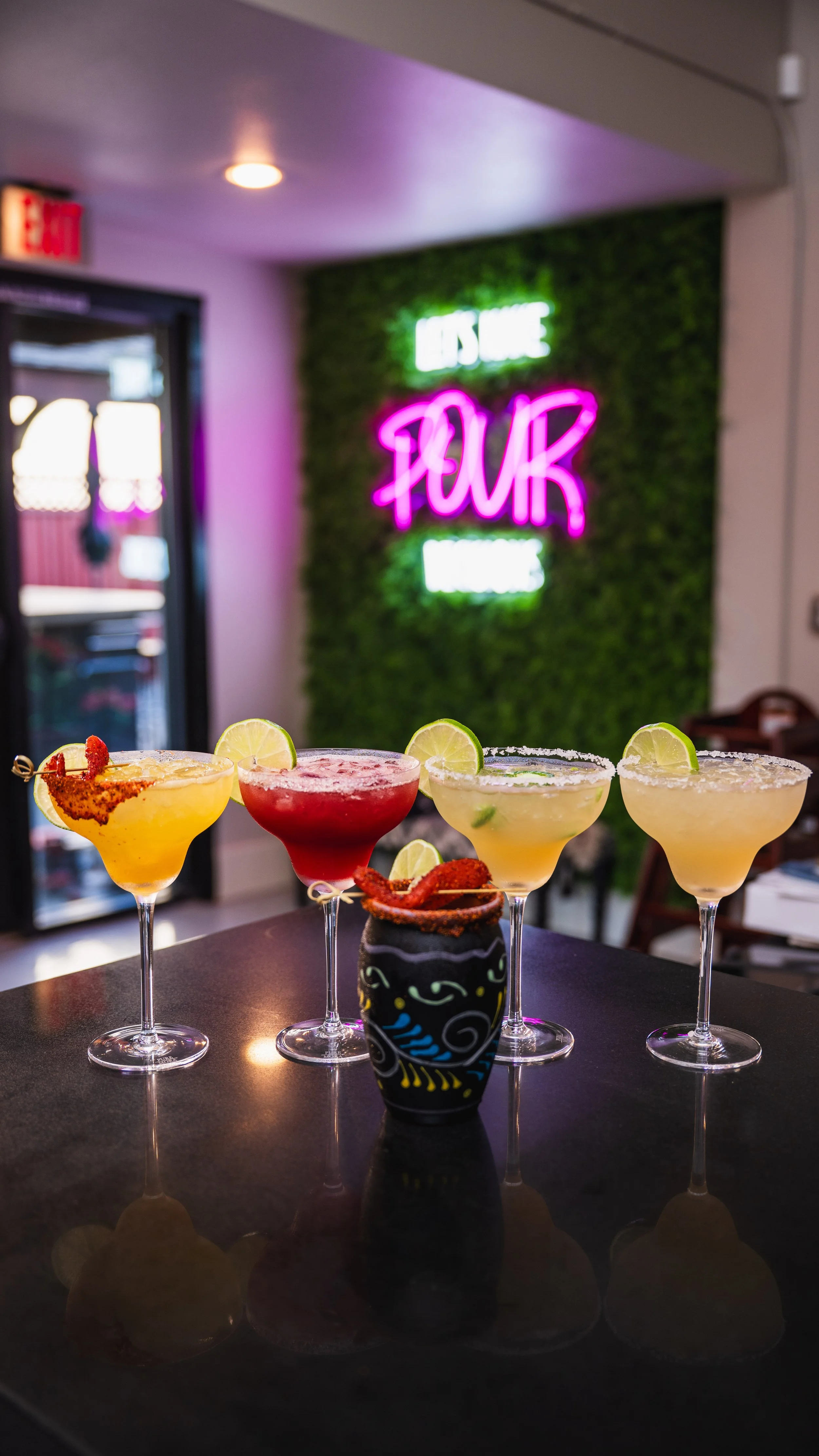 Five colorful cocktails with lime garnishes on a dark bar counter, with a neon sign reading 'FLOUR' and 'HOUSE' on a green wall in the background.