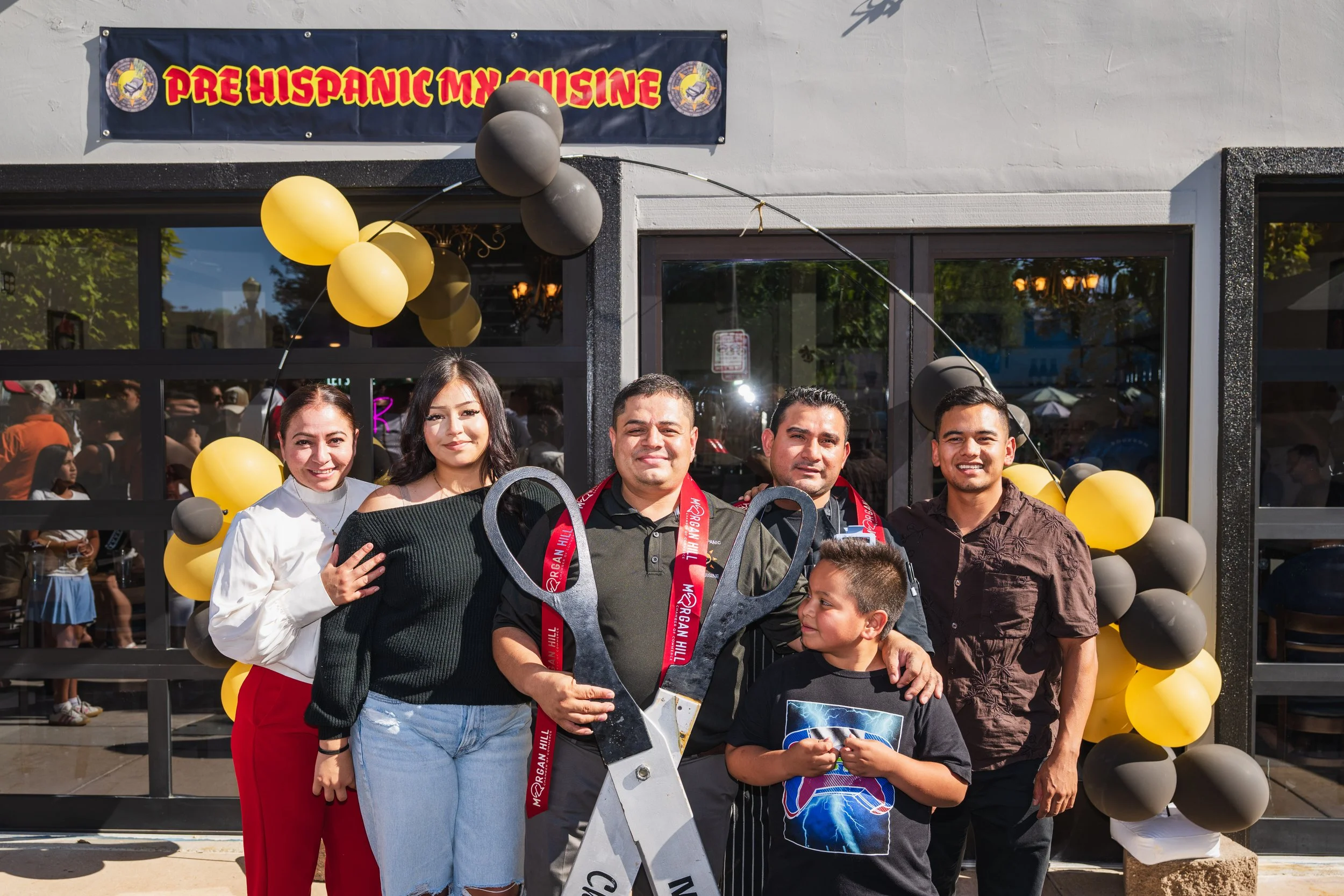Group of six people standing outside a building decorated with balloons, participating in a ribbon-cutting ceremony with oversized scissors. A banner above reads 'Pre Hispanic My Cuisine'.