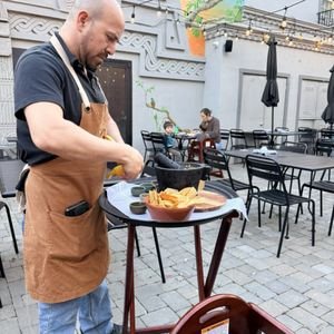 A man preparing food at an outdoor patio cafe with empty tables and chairs, and a white wall with decorative elements.