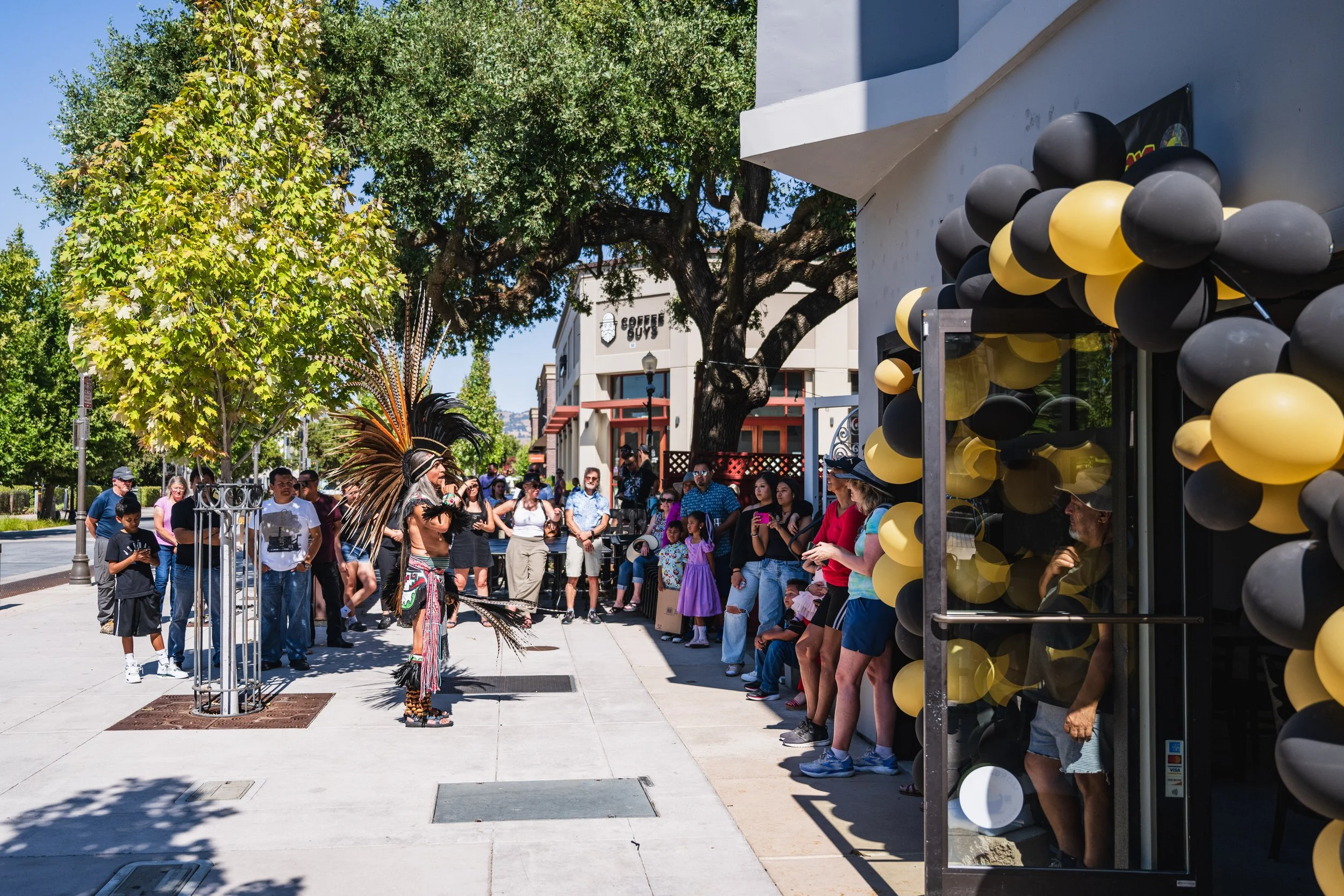 A street performer dressed in traditional indigenous costume with feathered headdress performs in front of a diverse crowd of onlookers gathered on the sidewalk. The scene is set on a sunny day with trees and storefronts visible in the background, and black and yellow balloons decorate the entrance of a nearby building.