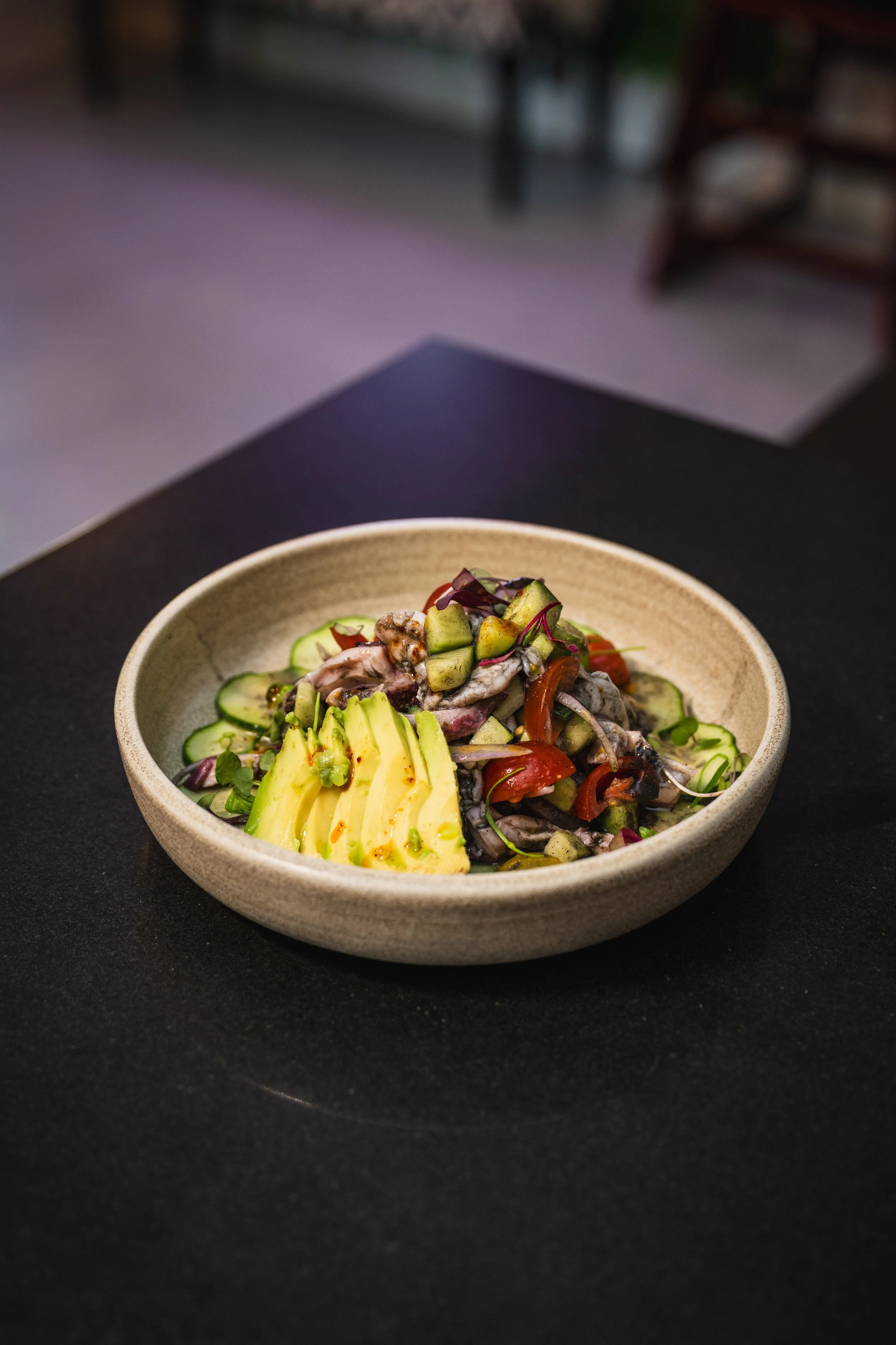 A bowl of mixed fresh vegetables with sliced avocado and cucumber on a dark table.