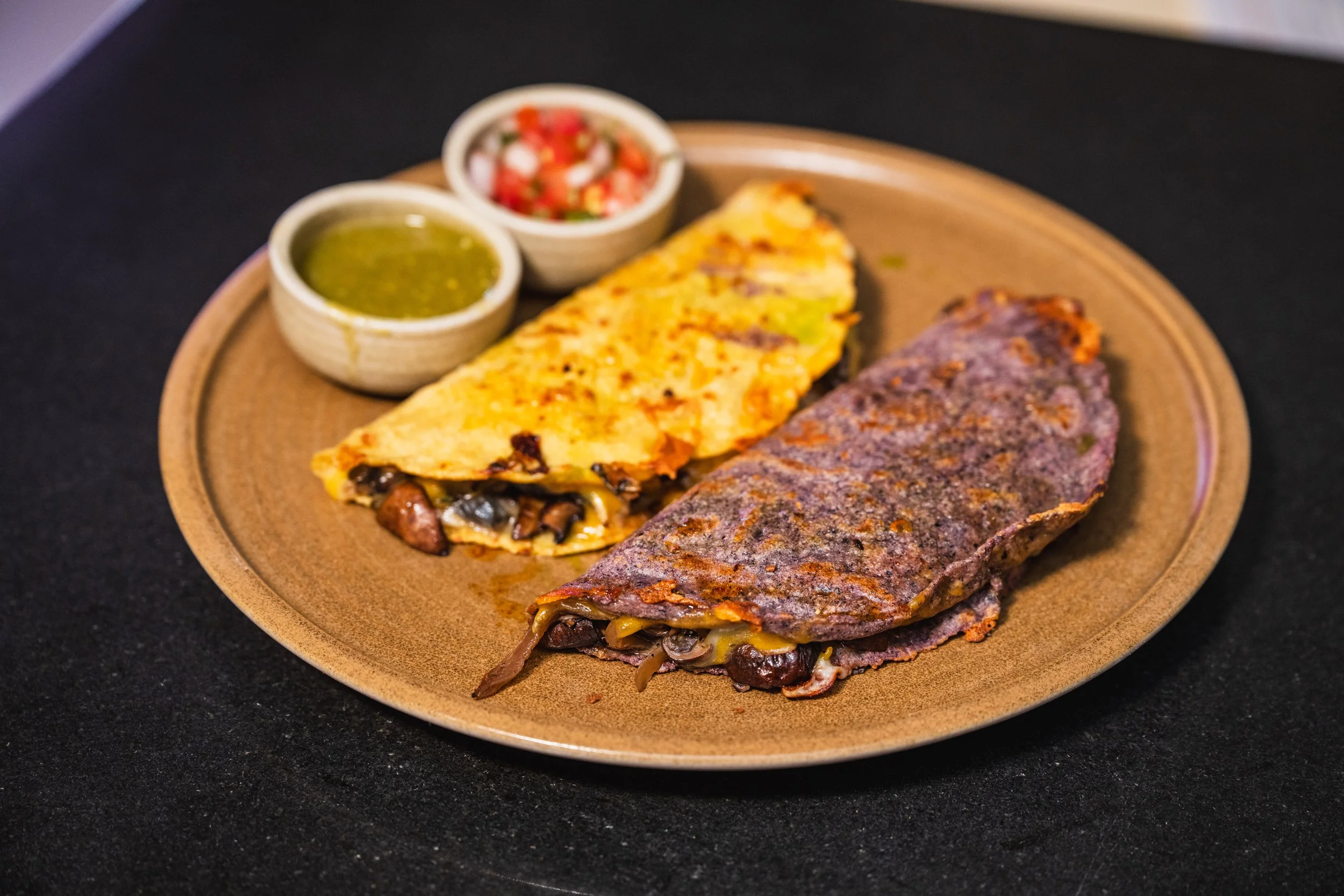 Plate with two beef fajitas, one with cheese, mushrooms, and peppers, and the other with peppers and vegetables. Accompanying are two small bowls of salsa and green sauce.
