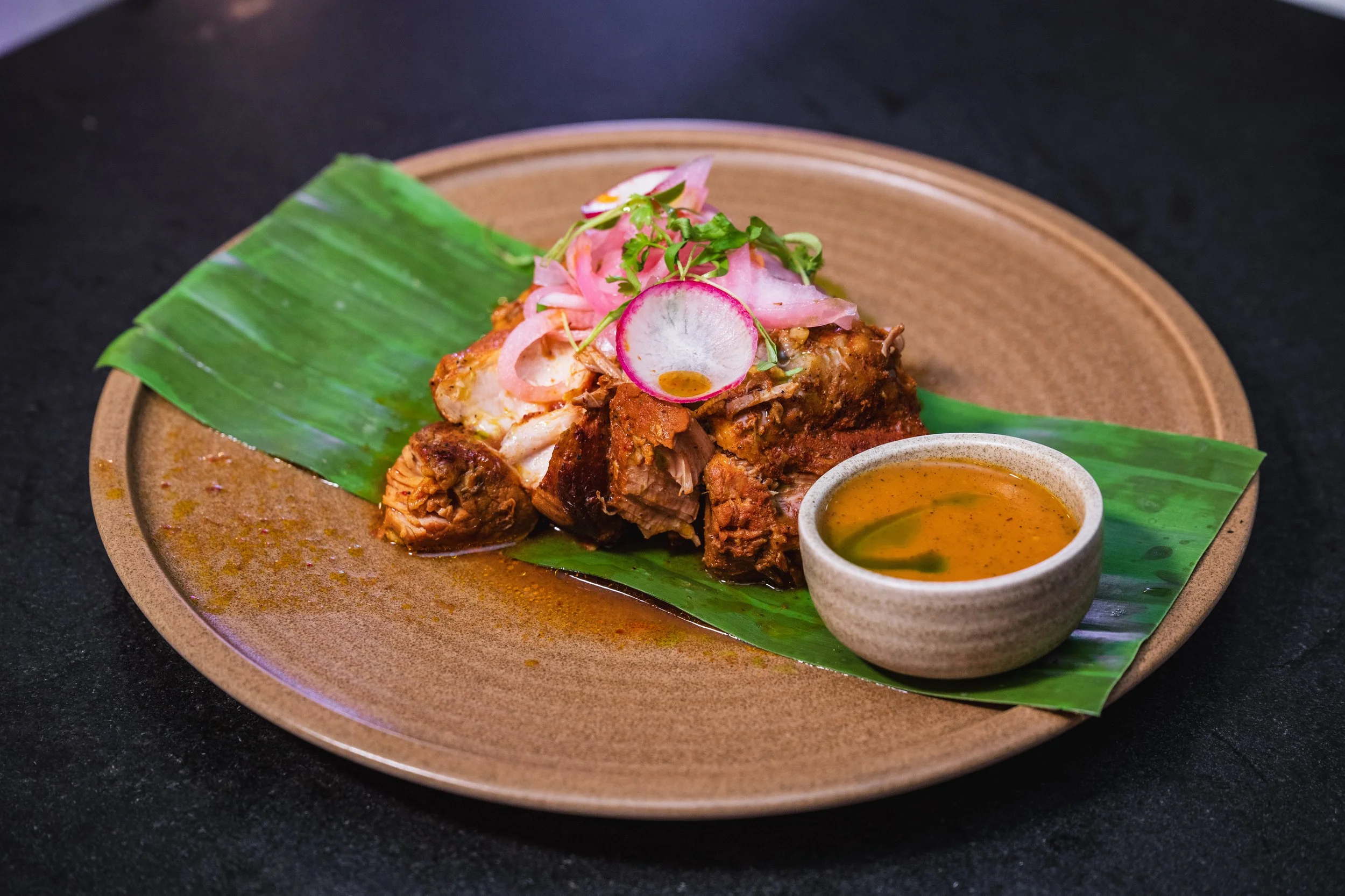 A plate of Mexican pulled pork with pickled red onions, radish slices, and microgreens, served on a banana leaf with a small bowl of sauce on the side.