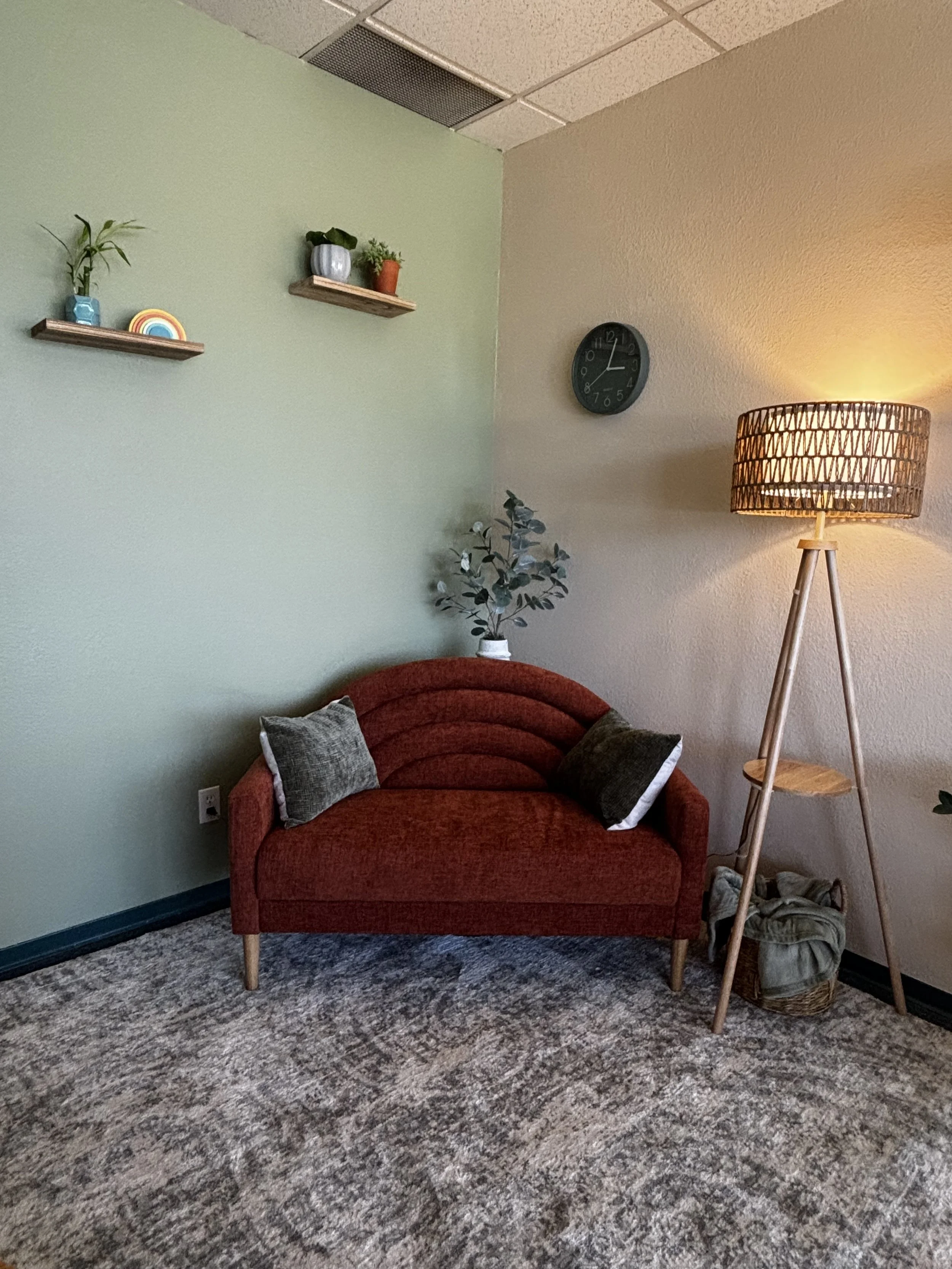A cozy office corner with a small red velvet sofa, decorative pillows, a potted plant, a floor lamp with a woven lampshade, and wall decor including two floating shelves with small plants and a rainbow figure, along with a wall clock.